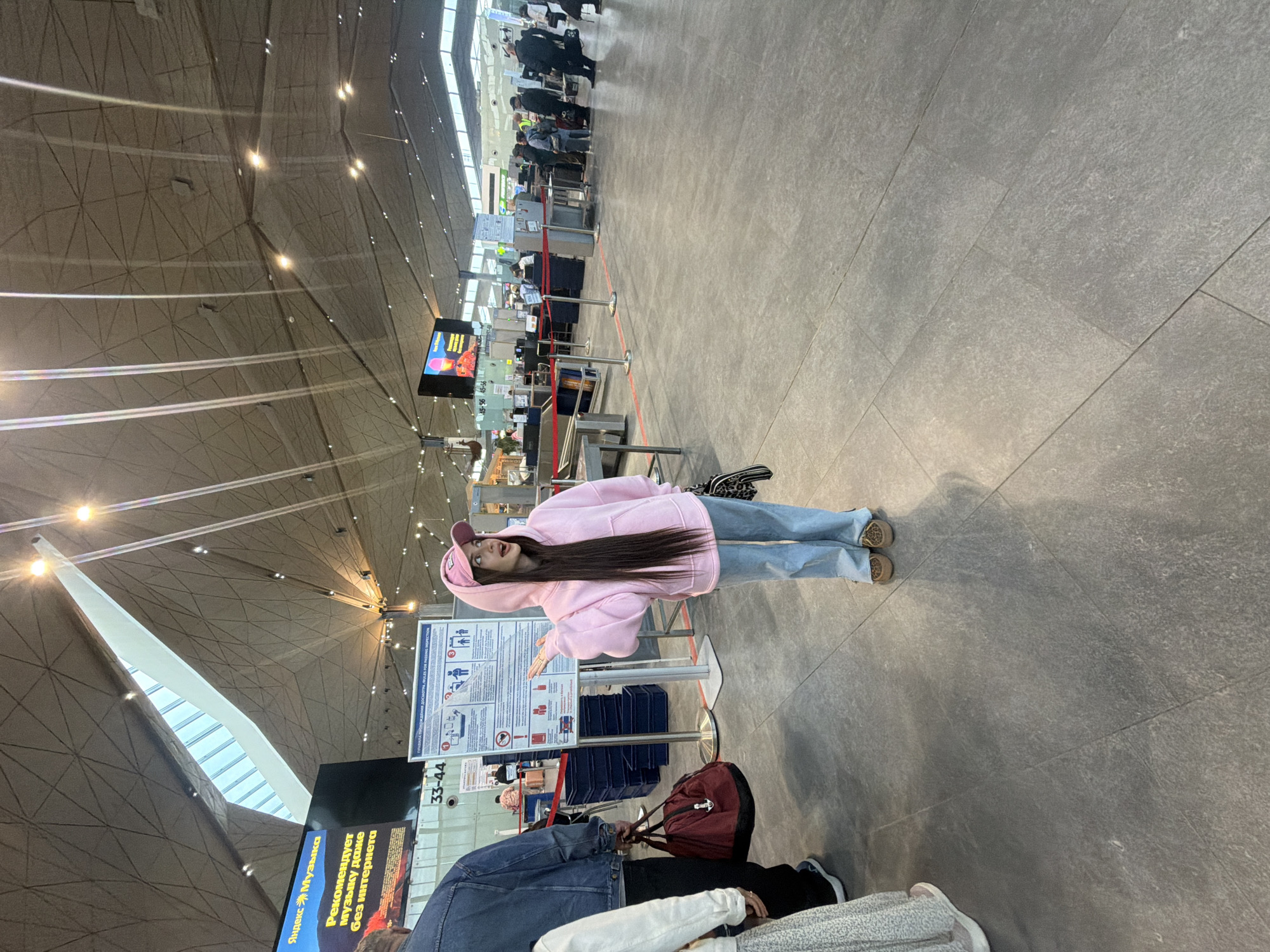The image shows an indoor scene likely within the Pulkovo Airport in Saint Petersburg, Russia. The architectural design of the airport's interior is evident, with a high, multifaceted ceiling structure. A young woman wearing a pink hoodie, a matching cap, and light blue jeans stands in the foreground. She appears to be interacting with a sign or poster, as suggested by the position of her arm. There are other people and luggage nearby, suggesting a typical airport environment. The flooring is tiled, and the lighting appears to be a mix of natural and artificial sources. Queues are forming at security or immigration points, with many people. A display screen with the number 33-44 is visible, likely marking the location.