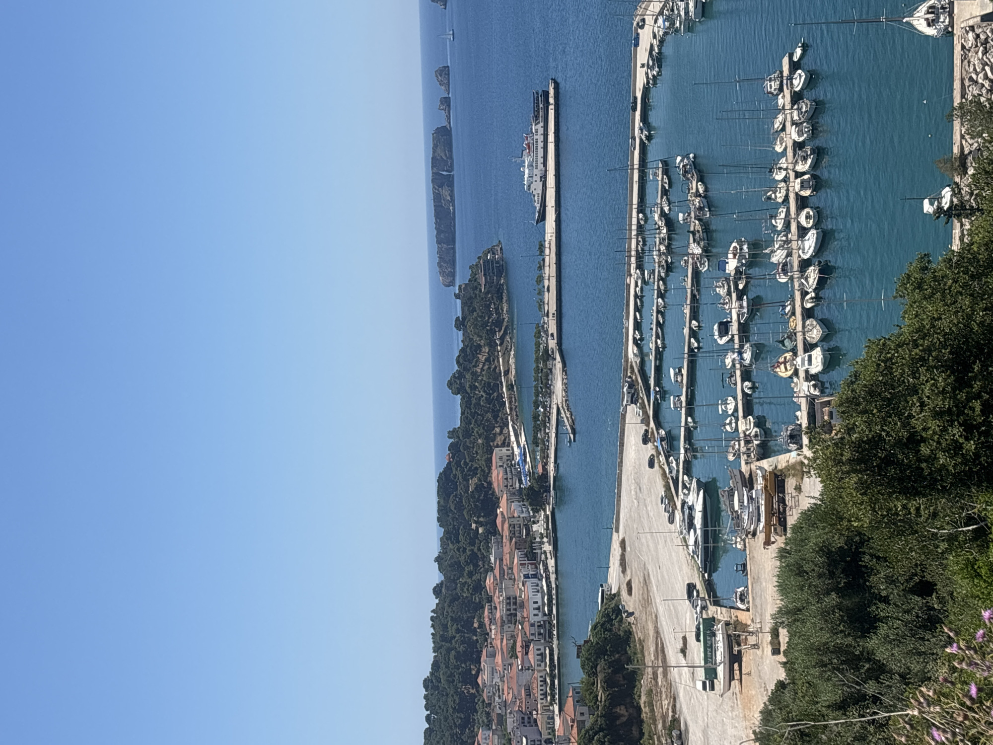 The image captures a coastal view of Pylos, Greece, featuring a marina filled with numerous sailboats. A large ferry is docked at a pier, with small islands visible in the distance. The town's buildings are nestled among lush green trees. The sky is clear blue.