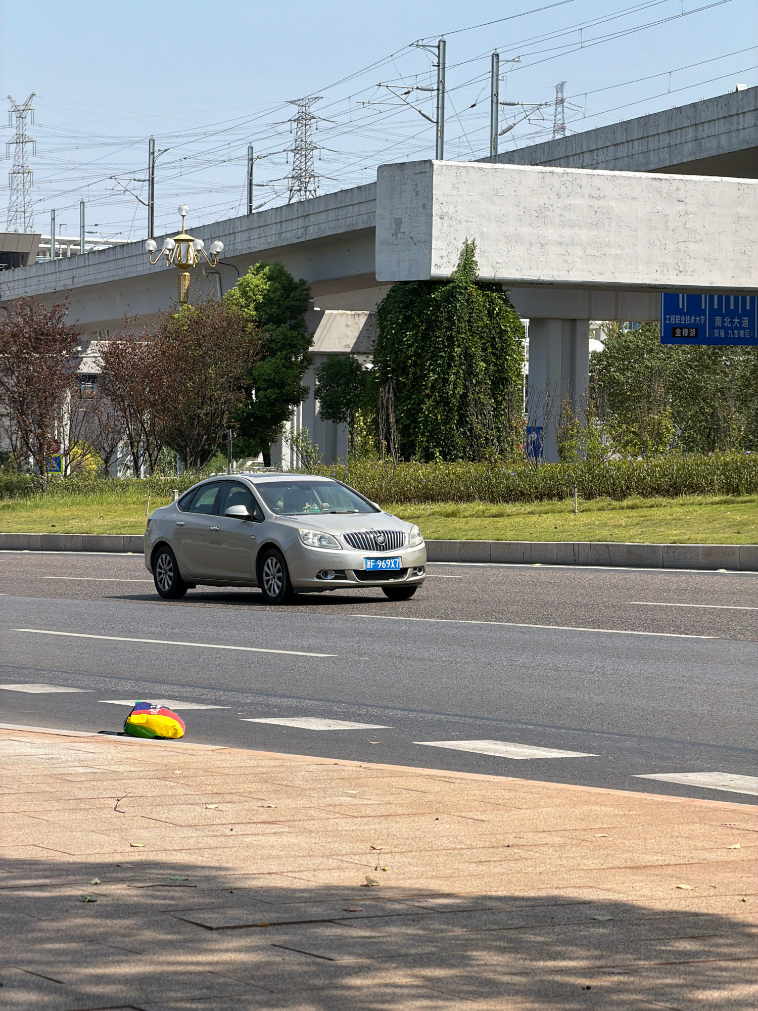 A silver four-door sedan is driving on a paved road in Jiangjin, China. The car has a license plate that reads "F-969X7". In the background, there is an elevated overpass with signs pointing to "Engineering Vocational and Technical University", "South-North Avenue", "Jinpu Lake", and "Shuanglong Jiulong District". There are also power lines and towers in the distance. On the sidewalk, a colorful object, possibly a deflated ball, lies on the ground. The weather appears to be clear and sunny.