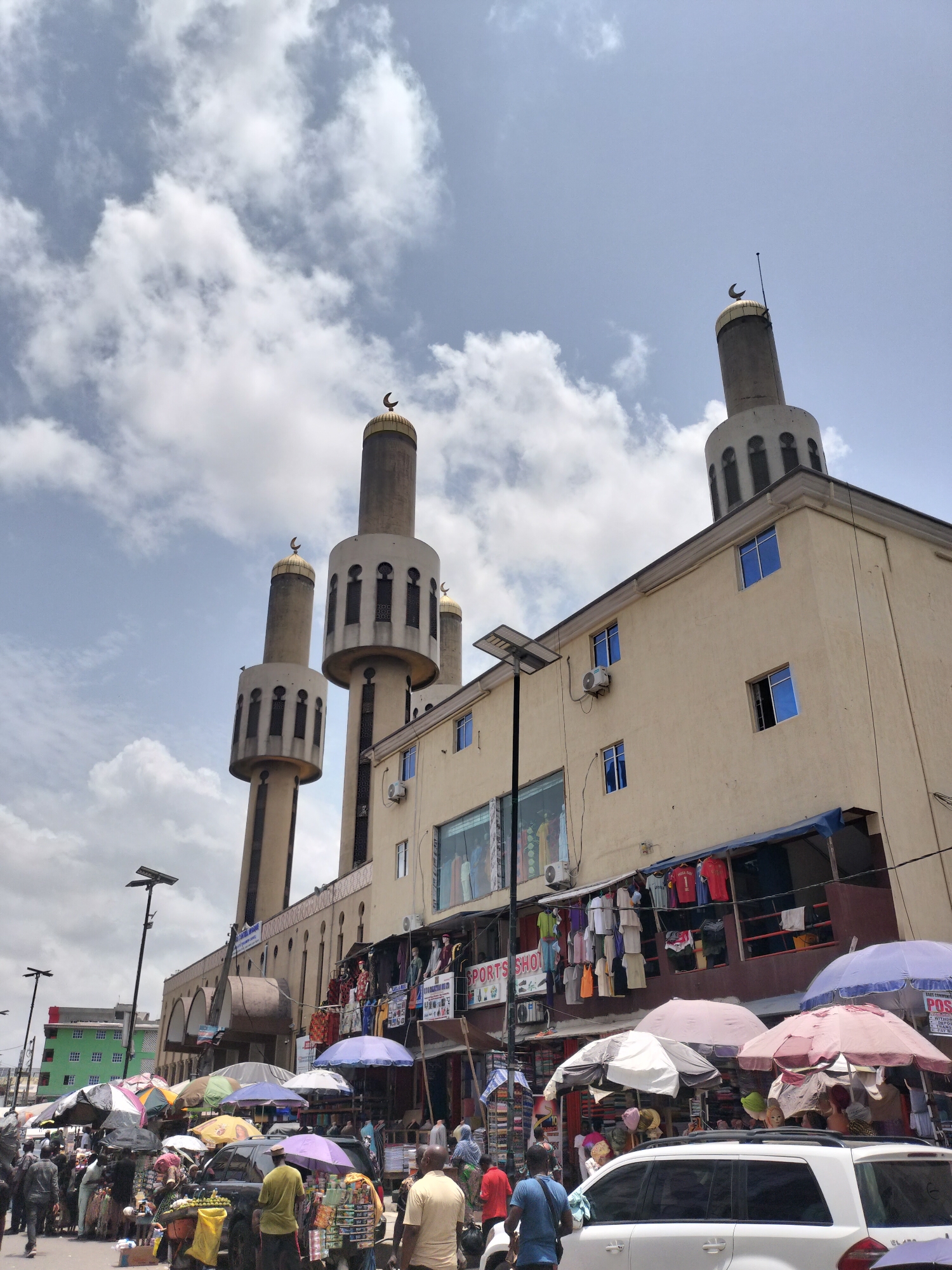 The image shows a mosque located on a busy street in Lagos, Nigeria. The mosque has four tall minarets topped with crescent moons. The beige-colored building is adjacent to a street lined with market stalls covered by umbrellas, where people are selling goods. Pedestrians and vehicles navigate the bustling scene. The sky is overcast with scattered clouds. A sports shop with clothes hanging is visible on the building next to the mosque.
