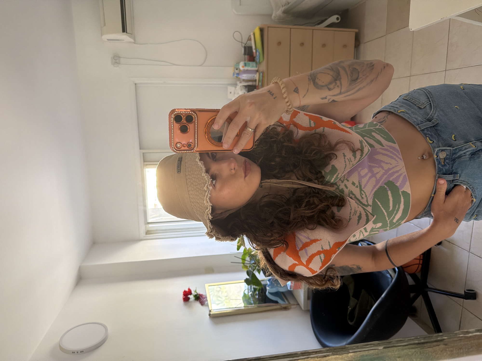 A woman is taking a selfie in front of a mirror, likely in her residence. She's wearing a patterned crop top and denim shorts, with a bucket hat on her head, holding an orange cellphone. Her left arm has tattoos. Behind her, in the reflection, are a window, a plant, and a black chair. The location appears to be an interior setting with tiled floors and white walls, possibly a room in a home. The location information given isn't visually evident and so cannot be definitively stated from the image.