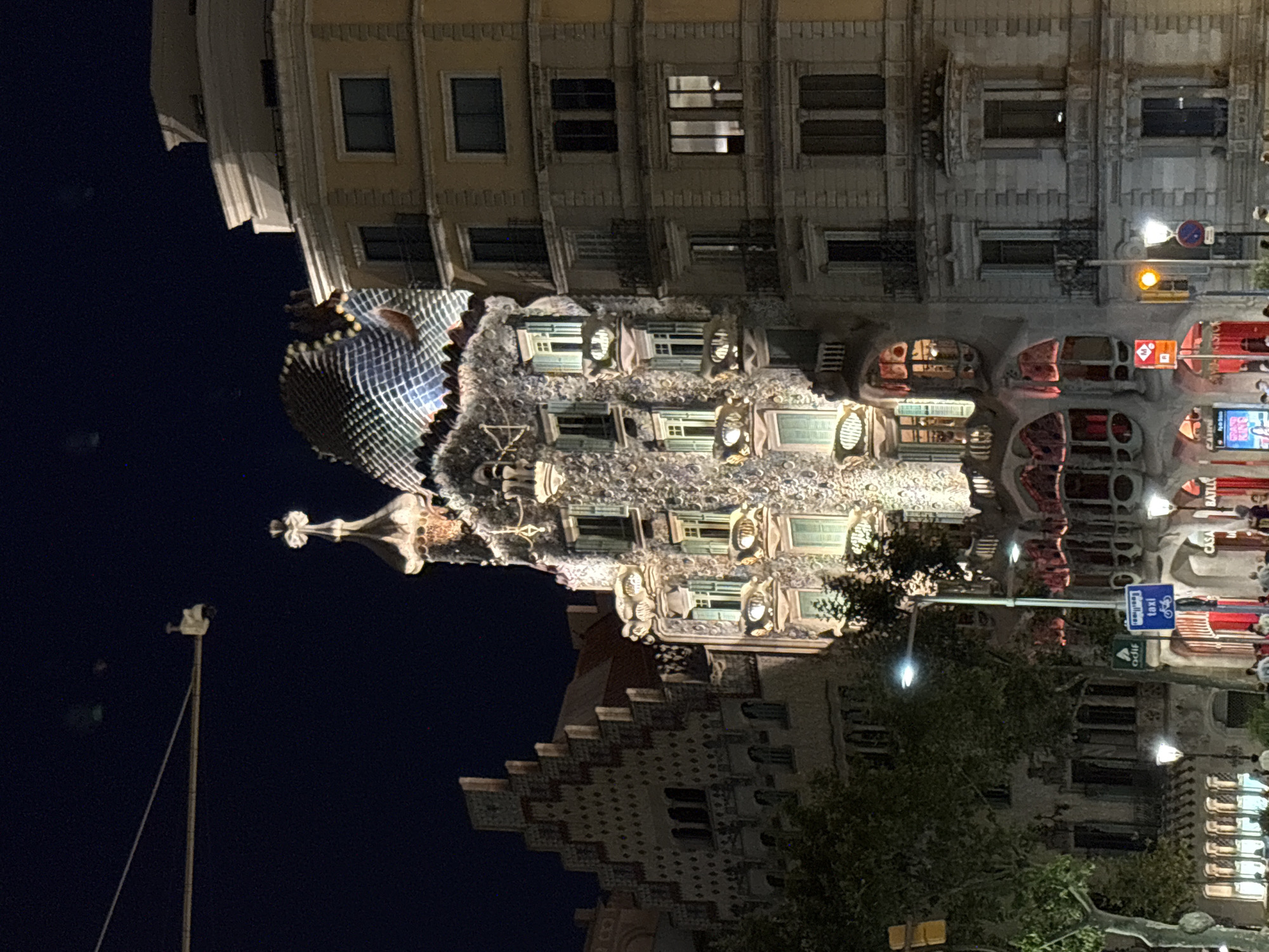The image shows Casa Batlló, a famous building designed by Antoni Gaudí, located on Passeig de Gràcia in Barcelona, Spain. It is night, and the building is illuminated, highlighting its unique, organic architecture. The facade features an array of colorful, mosaic-like tiles, irregular balconies resembling skulls, and a roof that resembles the scales of a dragon. The building is surrounded by other architectural structures, including a more conventional apartment building to its right. In the foreground, trees and streetlights are visible, along with a taxi sign and some people walking on the sidewalk. The sky is dark, indicating it is nighttime.