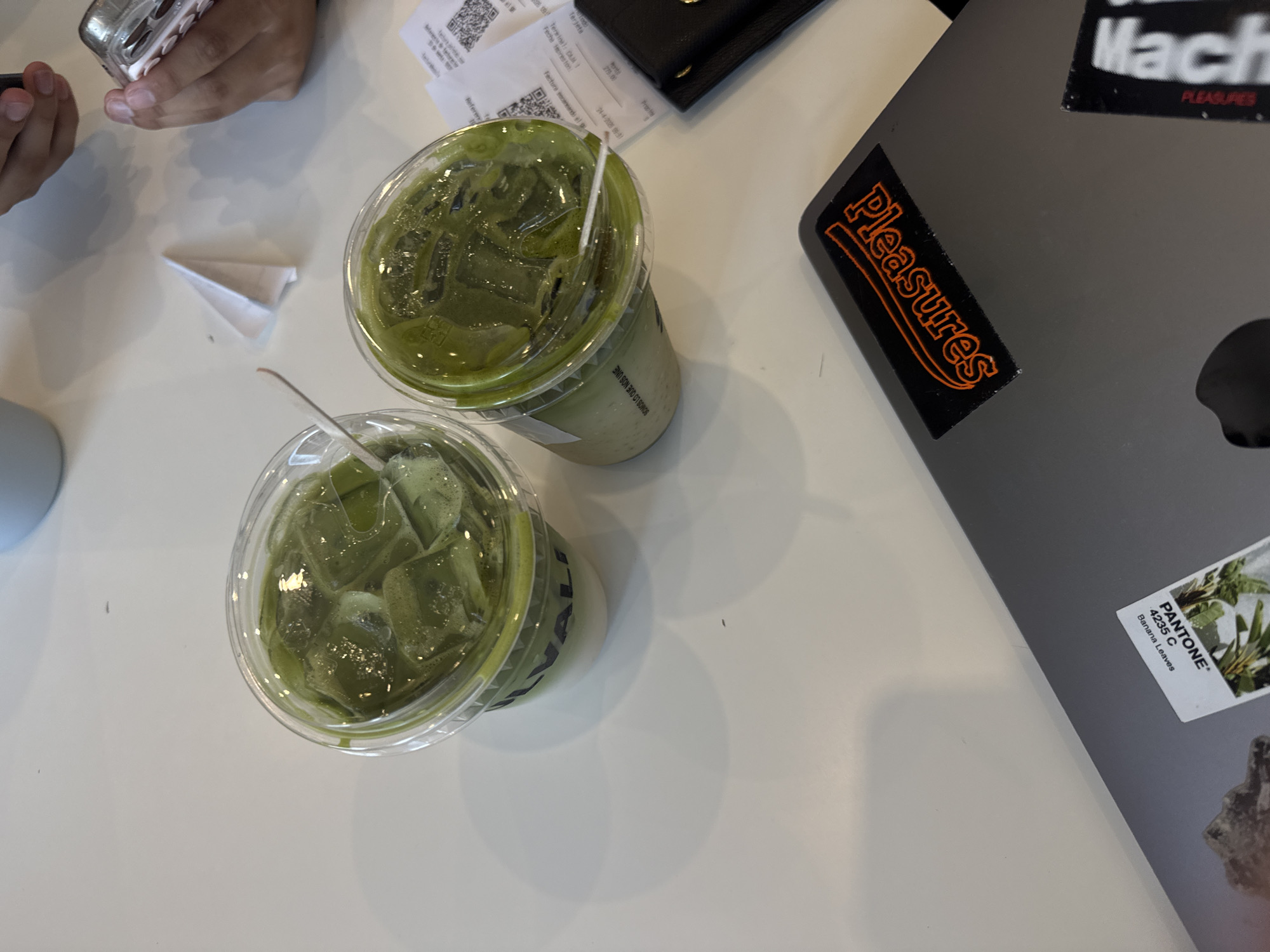 An overhead shot captures a bright, modern scene on a white tabletop, likely in a cafe or co-working space in Puebla, Mexico. The primary subjects are two iced green beverages, resembling matcha lattes, in clear plastic cups filled with ice and topped with froth. One cup prominently displays the Spanish phrase "SOMOS LO QUE NOS UNE" (We are what unites us) and part of a brand name "ALI".
In the upper left, a person's hands are visible, holding a mobile phone with a metallic case. Near the hands, a small white paper airplane rests on the table. To the right of the drinks, a dark-colored laptop, possibly a MacBook, is present, adorned with several stickers. One prominent sticker features the word "Pleasures" in orange text on a black background. Another partially visible sticker shows "Mach" and "Pleasures" in black text, while a third displays "PANTONE® 4235 C Banana Leaves" alongside an image of banana leaves.
Behind the drinks, several paper receipts are scattered, one showing a date and time of "24-4-202X 09:31" (indicating a morning setting) and monetary value "279.00". The receipts contain Spanish text such as "Fecha Emision," "Terminal," "Referer," and "Factura escaneando el," which, along with the "SOMOS LO QUE NOS UNE" text, provides strong location cues consistent with Puebla, Mexico. A black wallet or small purse is also partially visible among the receipts. The activity appears to be casual, perhaps a morning meeting or work session, with drinks and a person engaged with their phone.