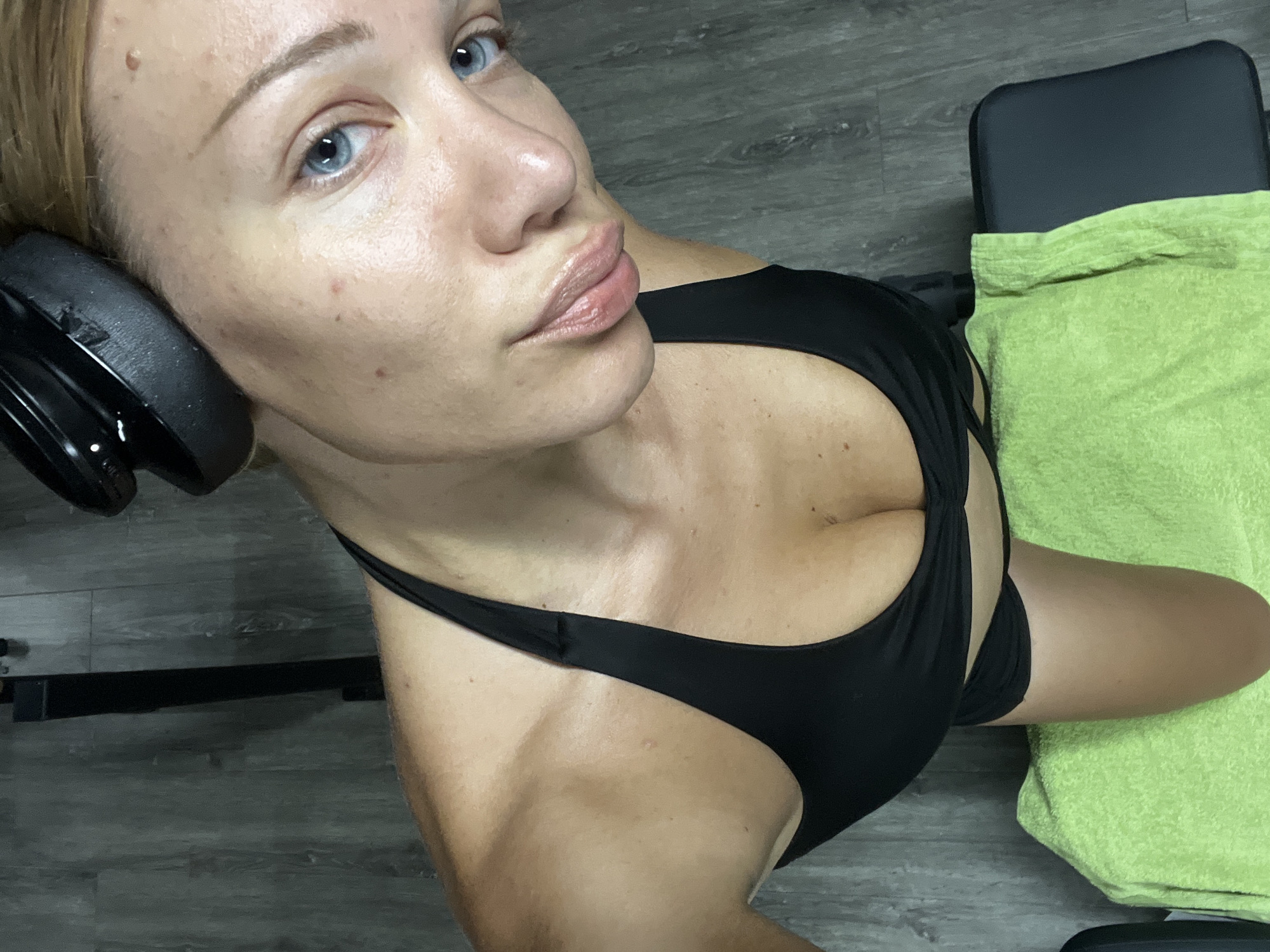 A young woman with blonde hair and blue eyes is taking a selfie in what appears to be a gym. She is wearing a black sports bra and black shorts. A bright green towel is laid out on a black bench in front of her. She is leaning forward, with a slight pout on her lips. Over-ear headphones are resting on top of her head. The background consists of a grey wooden floor and a black weight rack. The image shows the woman's face, shoulders, and upper torso. The lighting suggests artificial gym lighting. There is no indication of the specific location within Altötting, Germany.