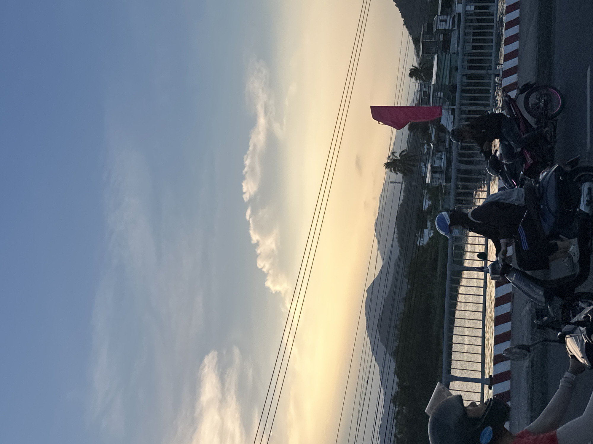 The image, taken in Nha Trang Ward, Vietnam, shows a sunset sky with clouds and power lines. Below, a scene on a bridge or elevated road is visible. Several motorbikes are parked along a concrete barrier with red and white stripes. People are standing and sitting near the motorbikes. In the background are houses and a mountain range. A red flag is visible in the distance.