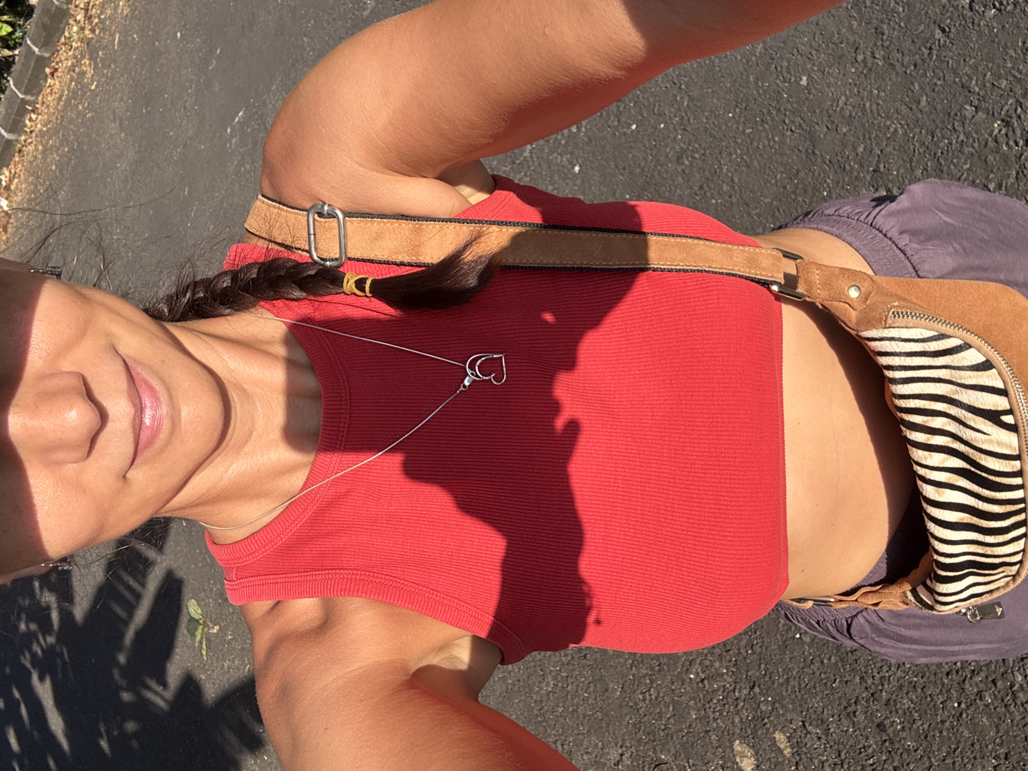 A woman with tanned skin stands on a paved road, her head and upper body facing the camera. She is wearing a red ribbed tank top and purple shorts. A brown leather bag with a zebra-striped section is slung across her torso. She has a dark braid in her hair, secured with a yellow band, and wears a delicate silver necklace with a heart-shaped pendant. Part of her face is in shadow, presumably cast by the bright sunlight. The location is Kuta Selatan, Indonesia, inferred from the submission instructions. The background is an asphalt road, partially shaded with trees.