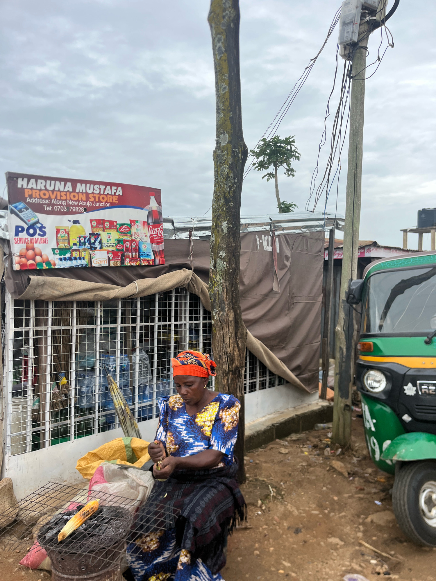 A woman, dressed in a blue and yellow patterned top and a dark patterned skirt with an orange headscarf, sits on the ground next to a charcoal grill with a piece of corn cooking. She is in front of a provision store with a sign that reads "HARUNA MUSTAFA PROVISION STORE" and an address that indicates it is located "Along New Abuja Junction". The store is stocked with various goods, including what appear to be bottles of drinks and eggs displayed on a shelf. A green and yellow auto-rickshaw is parked to the right of the scene. The environment is somewhat cluttered with debris on the ground and utility poles with numerous wires overhead. The sky is overcast. The scene appears to be a street-side vendor in Jos, Nigeria.