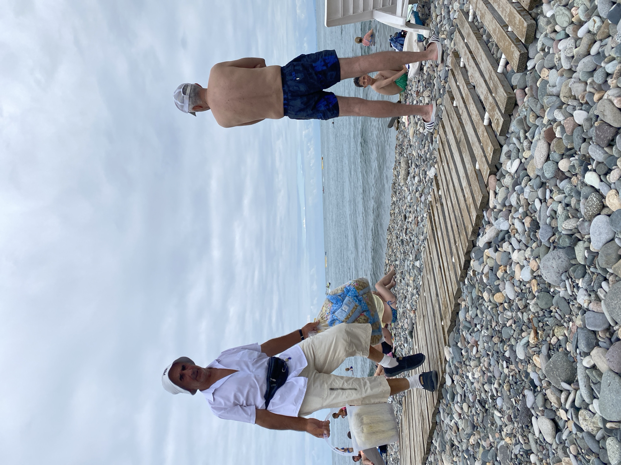 The image captures a scene on a pebbled beach in Batumi, Georgia, under an overcast sky. In the foreground, a man wearing a white bucket hat, a white short-sleeved shirt, beige capri pants, white socks, and dark slip-on shoes walks on a wooden boardwalk. He is carrying a clear plastic container with items inside in his left hand and a large, clear plastic bag filled with packaged goods in his right hand. A black fanny pack is worn around his waist. Behind him, another man, shirtless and wearing a white baseball cap and dark patterned swim trunks, stands facing away from the camera, his arms crossed. Further back, a young boy in green swim trunks sits on the pebbles. In the background, the calm sea stretches to the horizon, with a few small boats visible. Other beachgoers are scattered on the shore and in the water, some partially obscured. The environment is characterized by the grey pebbles covering the beach, the wooden boardwalk, and the tranquil ocean.