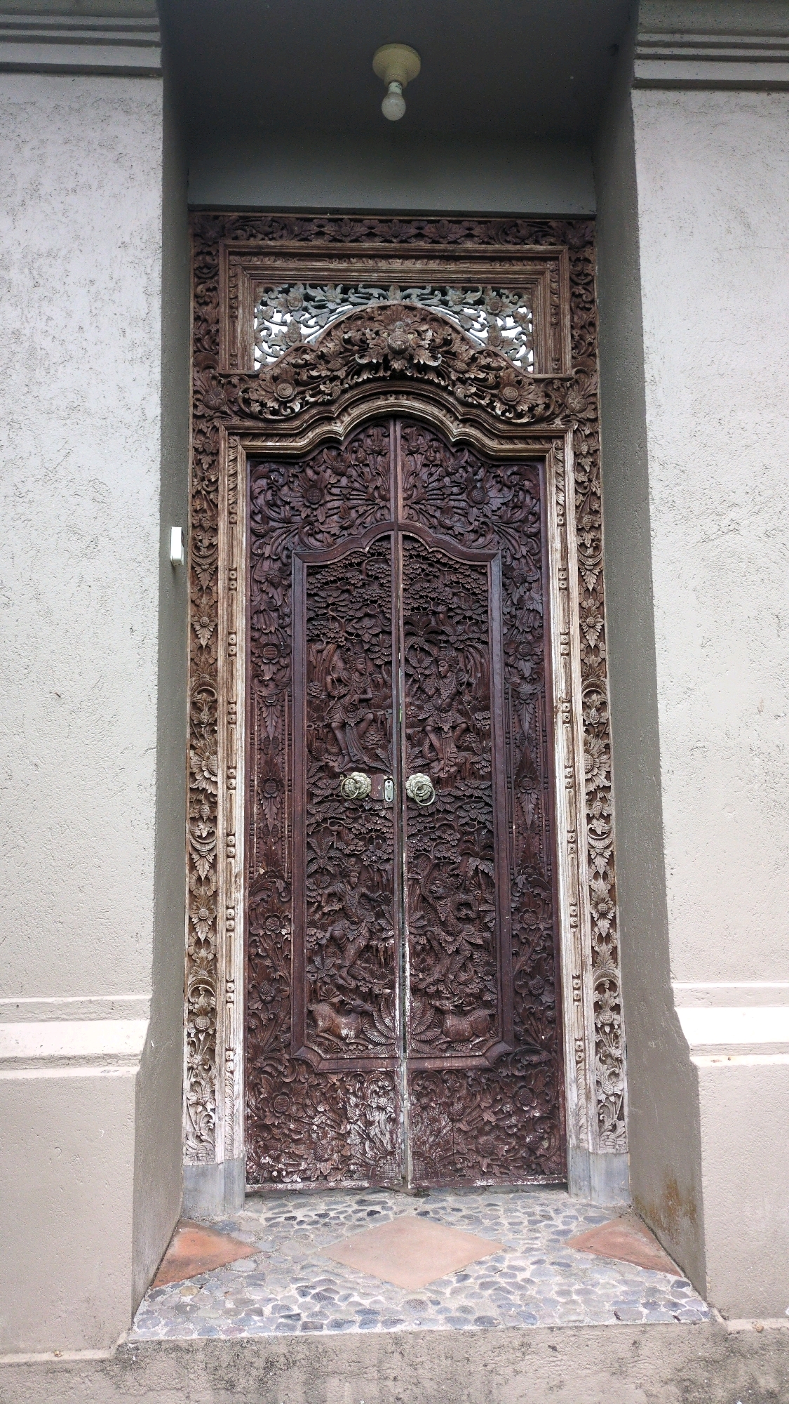 The image shows a highly ornate, dark wooden double door, intricately carved with figures and patterns, set within a textured, light-colored stone wall. The door features two brass door handles, positioned centrally. Above the door, a carved wooden frame surrounds a decorative grille, and a single light bulb hangs from the ceiling. The ground in front of the door is paved with small, multi-colored stones and two square tiles. The wall appears to be part of a building in Ubud District, Indonesia, given the distinctive architectural style and the lush, tropical environment implied by the elaborate carvings which often depict mythological scenes or local flora and fauna.
