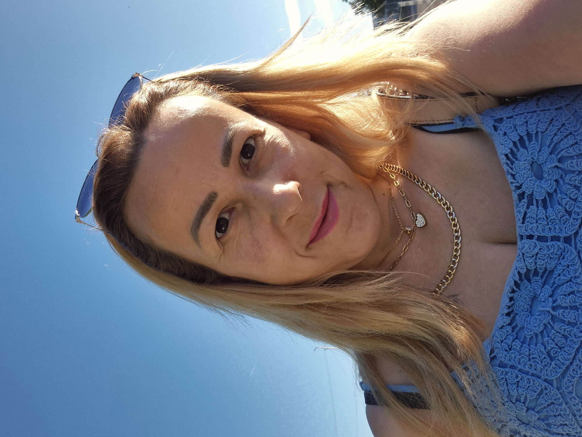 A woman with blonde hair and fair skin, wearing a blue crocheted top and gold necklaces, poses for a selfie outdoors against a clear blue sky. Sunglasses rest on top of her head. The lighting suggests it is daytime. The background is out of focus, showing a hint of a street and some distant trees. The image appears to have been taken on a sunny day.