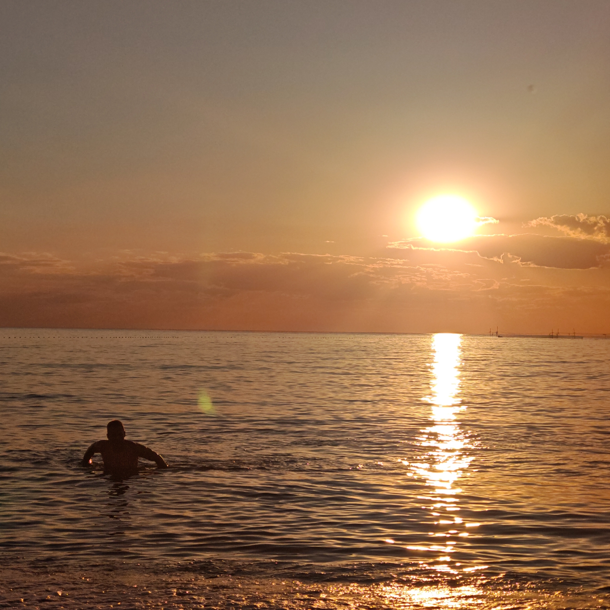 A man is swimming in the ocean during sunset. The sun is setting on the right side of the image, casting a golden glow over the water. The water is calm with gentle waves. The man is silhouetted against the sunset, and his body is partially submerged in the water. The sky is a gradient of orange and pink, with some clouds scattered across it. The scene appears to be a peaceful and serene moment.
