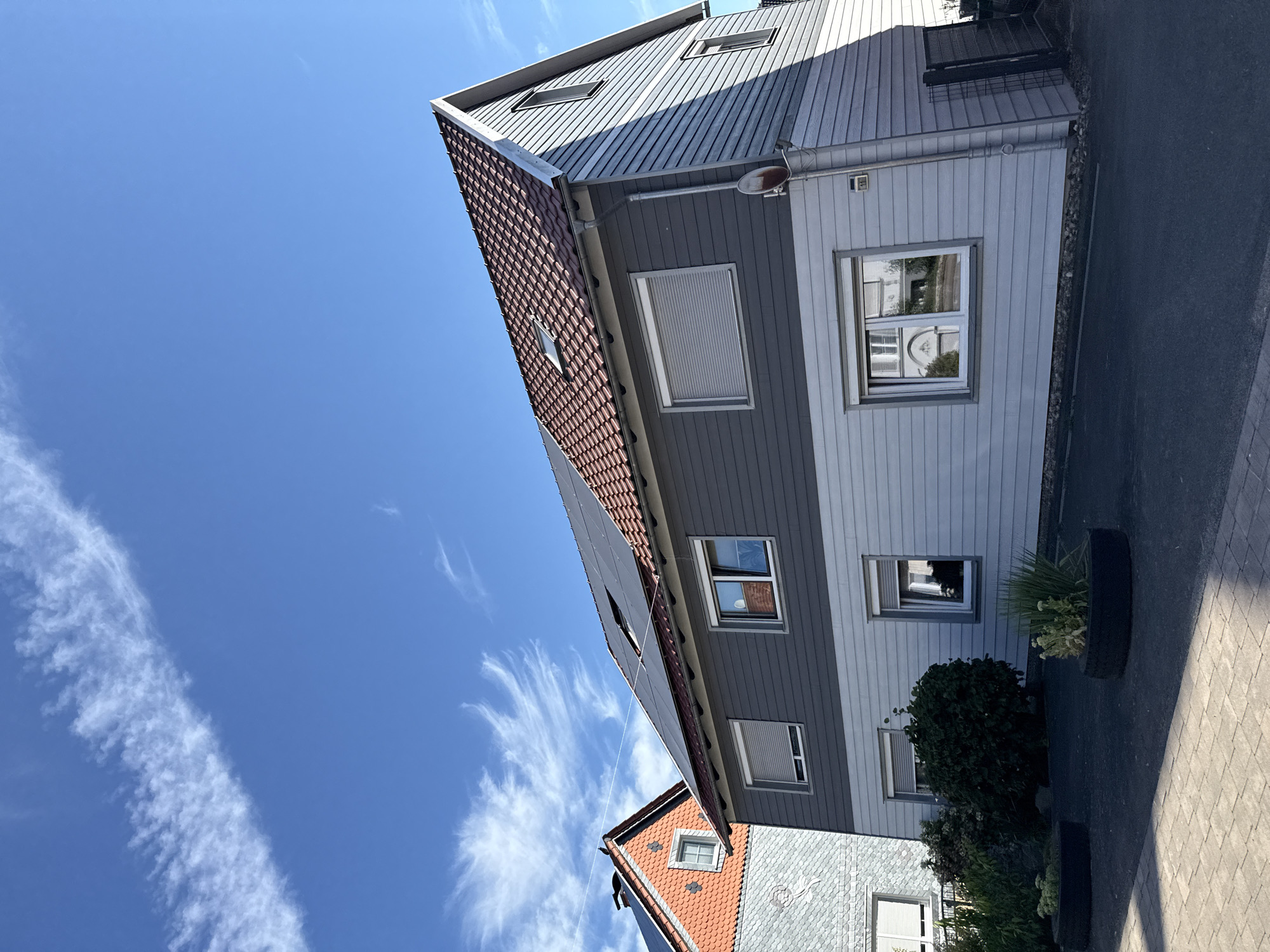 This is a full shot of a modern German house in Walkenried, Germany, with a dark grey upper story and a lighter grey lower story, topped with a red-tiled roof. Solar panels are installed on the roof. The house has multiple windows with grey shutters, some of which are closed. In the foreground, there is a paved driveway and some plants in tire planters. To the left, a portion of another house with an orange-tiled roof and grey slate siding is visible. The sky is a bright blue with some wispy white clouds.