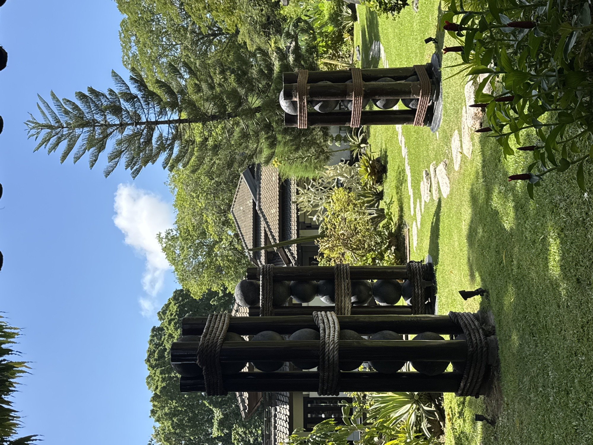 The image captures a garden scene in Kuta Selatan, Indonesia, featuring three decorative wooden structures lined with spherical stones. The structures are constructed from dark wood and bound together with thick, dark rope. They appear to be a form of garden art.
Behind these structures, a lawn is visible, dotted with stone pathways. Various trees and plants surround the lawn, including a tall evergreen tree with a distinctive branch pattern. Parts of a building are visible in the background, suggesting the garden is part of a larger property. The sky is clear and blue with a few white clouds. Red flowering plants are present on the right side of the frame. The lighting suggests it is daytime.