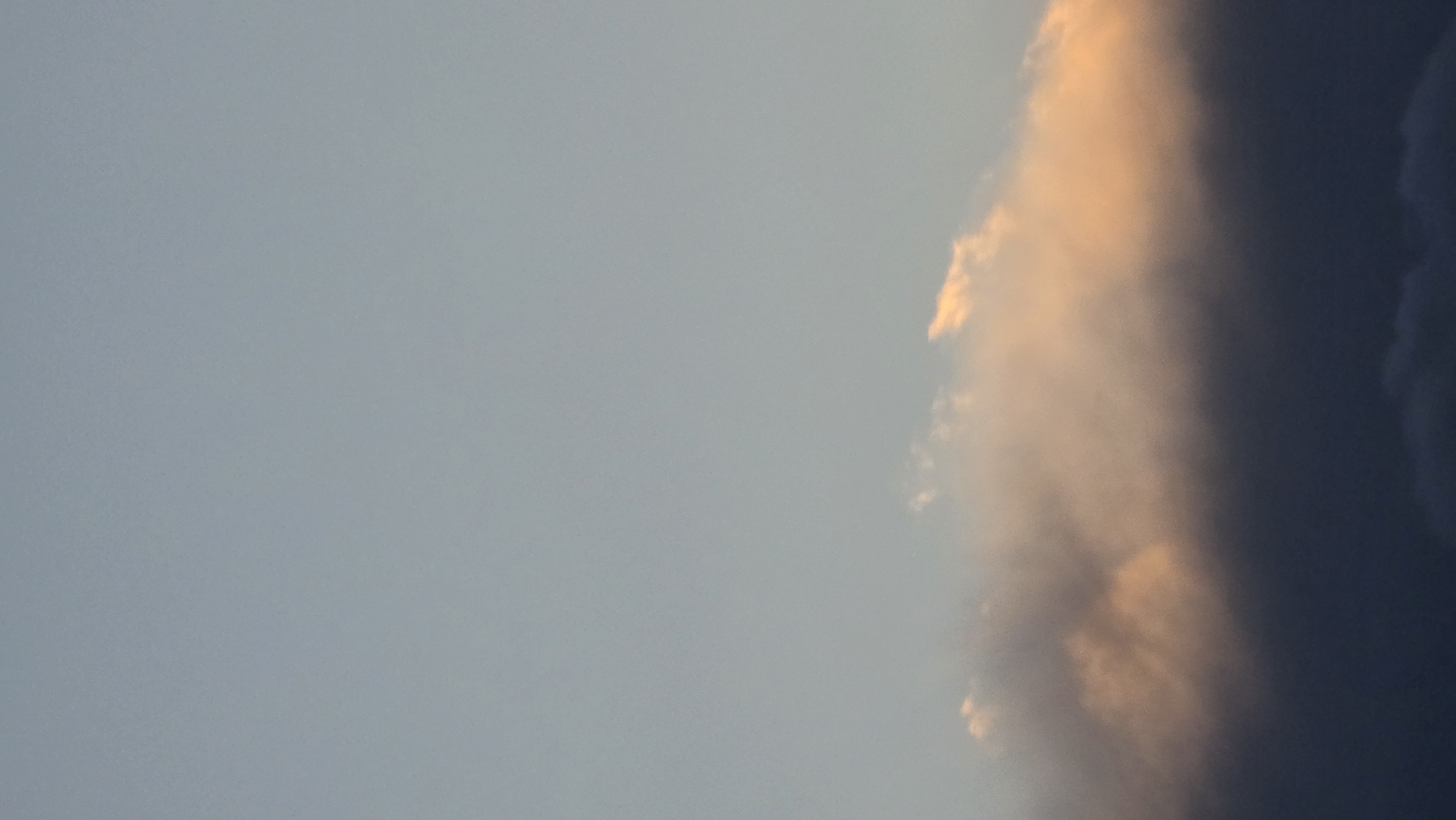 The image shows a portion of a cloudy sky, likely taken during sunset or sunrise. The left side of the frame is a light blue, transitioning towards a dark, almost black cloud formation on the right. Parts of the clouds are illuminated with a golden-orange hue. The image does not contain information to determine its location in Blagoevgrad, Bulgaria, or details about people, subjects, objects, actions, or interactions.