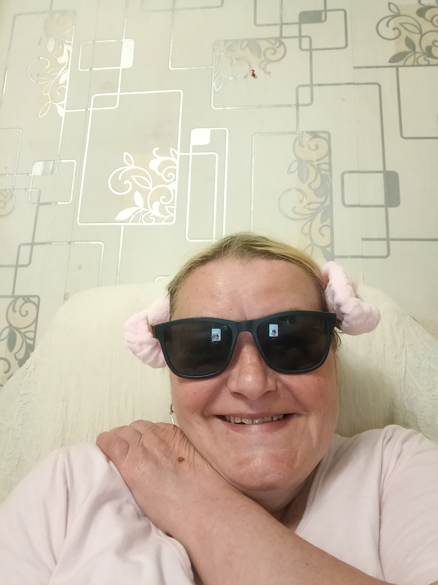 A medium shot shows a woman smiling and wearing sunglasses with pink fuzzy decorations on the sides, seemingly indoors. She is wearing a light pink shirt and rests her left arm across her shoulder. The background features a wall with a geometric pattern and floral designs. The photograph appears to have been taken in Красный Маяк, Russia, based on metadata.