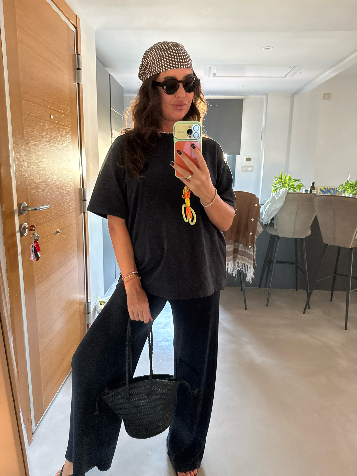 A woman stands indoors, taking a mirror selfie. She is wearing a black t-shirt, black wide-leg pants, and a patterned headscarf. She has on sunglasses and is holding a black woven basket purse in her left hand. The background shows a doorway on the left, and a room with stools and plants on the right. The scene appears to be in a residential setting.