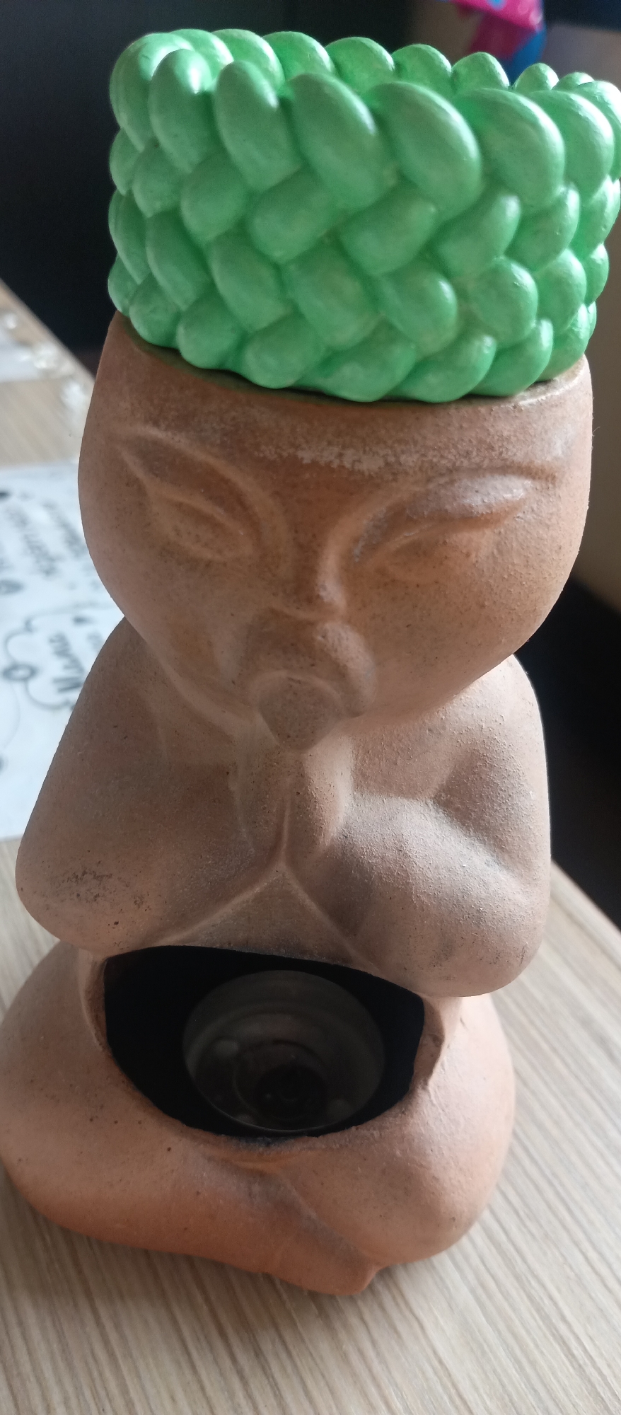 The image shows a terracotta statue of a figure with a stylized face and hands clasped in front of its chest. A green, braided-looking object sits atop the figure's head. The statue has an opening in the lower portion, revealing a dark interior with a visible bulb. The background is blurred, but the statue appears to be on a wooden surface. The location is Unknown, Unknown.