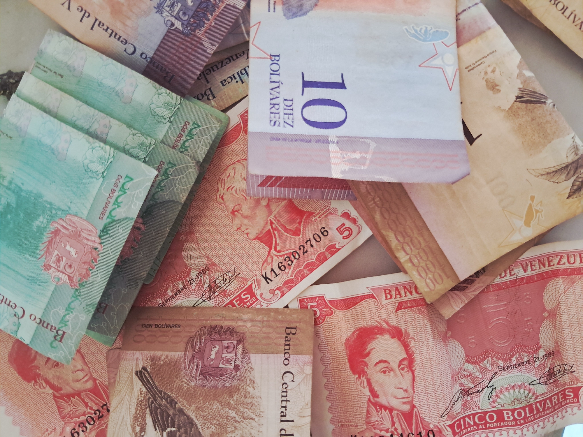 The image shows a collection of Venezuelan Bolivar banknotes of various denominations. The banknotes feature portraits of historical figures and national symbols. Denominations visible include two, five, ten, and one hundred Bolivares. The banknotes appear to be arranged haphazardly on a flat surface. There is no specific information about the location within Juan Griego, Venezuela, nor any indication of people, actions, or interactions in the scene.