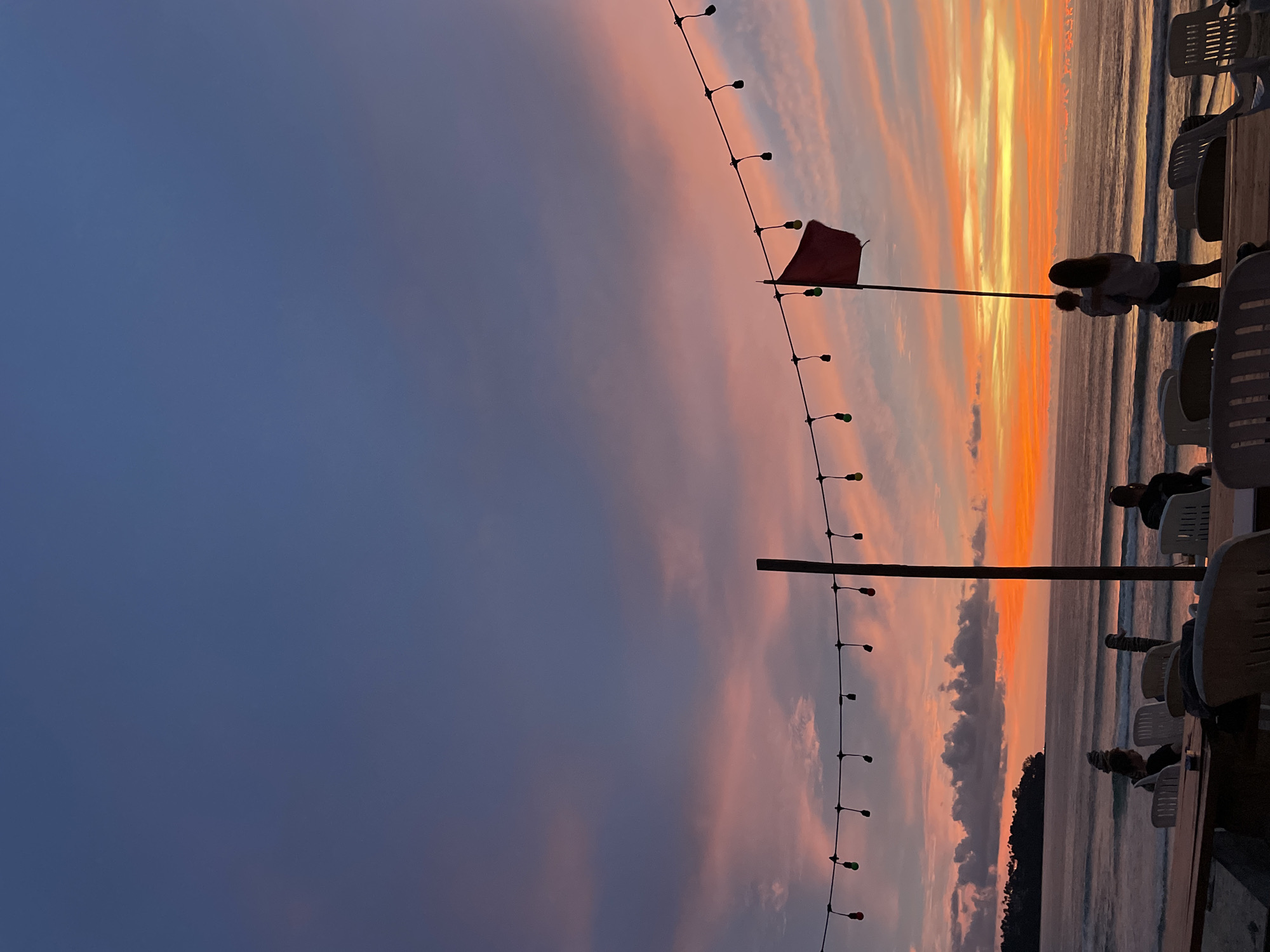 A vibrant sunset paints the sky over a beach in Choeng Thale, Thailand. The sky transitions from a deep indigo at the top to hues of pink, orange, and yellow near the horizon, with wispy clouds streaking across. A string of decorative lights with visible bulbs stretches across the mid-section of the image, casting a subtle glow. A tall, dark pole stands prominently in the foreground, topped with a red flag that flutters in the breeze. To the right of the pole, a woman holds a child, both silhouetted against the colorful sky, facing the ocean.
The sea is calm with gentle waves rolling towards the shore. In the distance, a tree-covered headland juts out into the water on the left side of the frame. The foreground reveals an outdoor seating area with white plastic chairs and tables, suggesting a restaurant or cafe. Several other people are seated at these tables, also appearing as silhouettes, enjoying the sunset view. The overall atmosphere is serene and picturesque, capturing the beauty of a tropical evening.