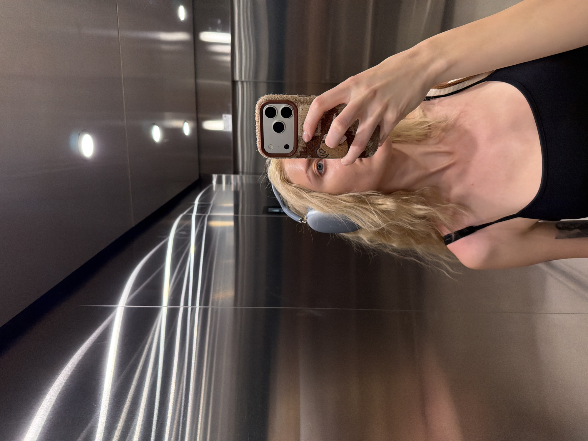 A woman is taking a selfie in an elevator with stainless steel walls. She is holding a brown phone in front of her face, partially obscuring it. She has blonde hair and is wearing gray headphones and a black top. The elevator has vertical light fixtures on the left wall. The location of the city of Belgrade, Serbia is not able to be determined in this image.