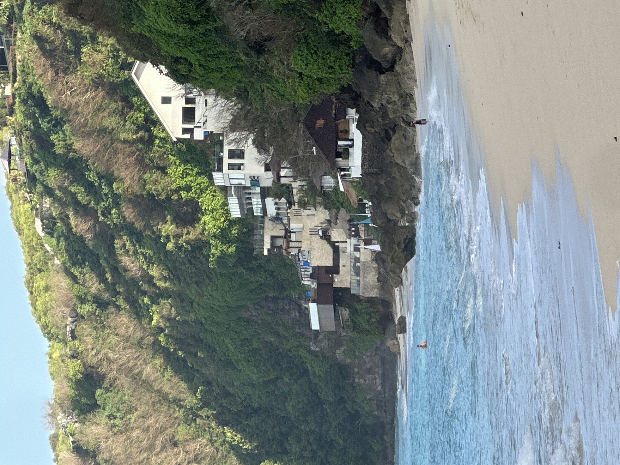 The image shows a coastal scene in Kuta Selatan, Indonesia, featuring a sandy beach with clear turquoise water and gentle waves. Several white buildings are built into the lush green hillside that rises steeply from the shore. These structures appear to be modern villas or resorts, characterized by multiple levels, balconies, and large windows. At the base of the cliff, a few smaller, darker-roofed buildings are nestled among the rocks. Two individuals are visible: one person is standing in the shallow water on the left side of the frame, and another is walking on the wet sand near the shoreline on the right. The sky is clear and light blue, suggesting a sunny day. The vegetation on the hillside is dense and varied, with patches of both green foliage and drier, more sparse growth.