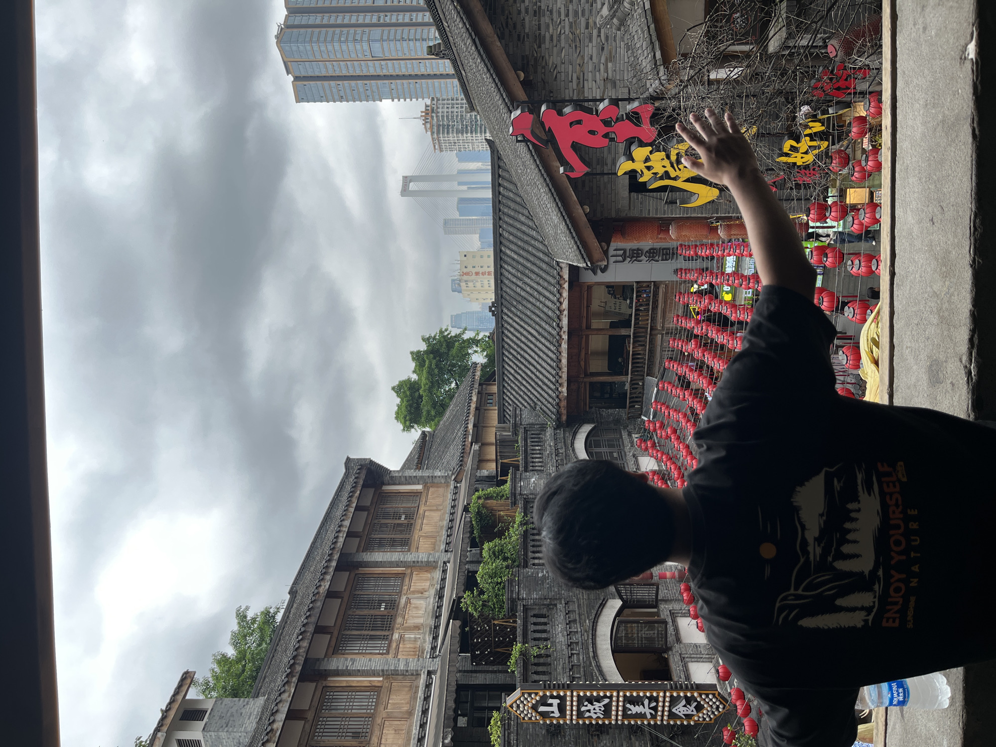 A medium shot captures the rear view of a person with dark hair, wearing a black t-shirt with a graphic print featuring trees, a mountain, and the text "ENJOY YOURSELF - SUNSHINE NATURE", as they extend a hand toward a display of red lanterns and oriental signage in red and yellow script. The individual is positioned within a shaded, seemingly interior space, offering a framed view of an urban landscape.
The architecture visible in the background features traditional Chinese building designs characterized by sloping tiled roofs, multiple stories, and wooden lattice windows. Modern skyscrapers rise in the distance, juxtaposing the old and the new within the city's skyline. The sky is overcast and grey. The location is in the city of Yuzhong District, China.