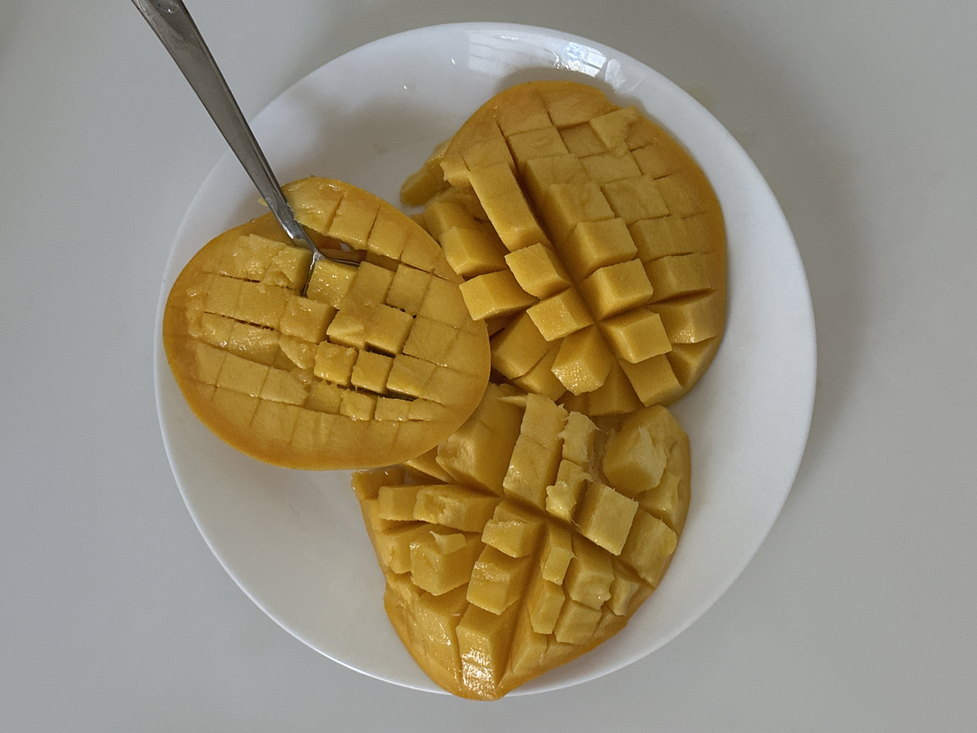 The image depicts a white plate containing three halves of a mango, cut into a grid pattern, with a silver fork inserted into one of the mango halves. The mangoes are yellow-orange in color, indicating ripeness. The setting appears to be indoors on a plain white surface. No people are visible. There is no indication of a location in Vrindavan, India.