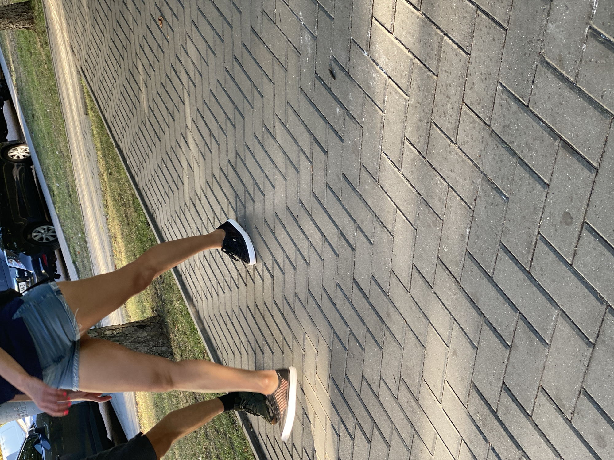 The image depicts a section of a paved pedestrian walkway with a herringbone pattern of gray bricks, likely in Chișinău, Moldova. The walkway is bordered by a strip of grass and, in the background, a road with parked cars. Sunlight casts long shadows across the brickwork, indicating it is either early morning or late afternoon.
In the foreground, two individuals are walking. The person on the left, whose lower legs and feet are visible, is wearing dark athletic shoes and black socks. They are walking on the grass verge next to the walkway. The person on the right, whose legs and feet are also visible, is wearing denim shorts and black sneakers with white soles. They are walking on the brick walkway. The overall impression is of a casual outdoor scene, potentially in a park or on a tree-lined street.