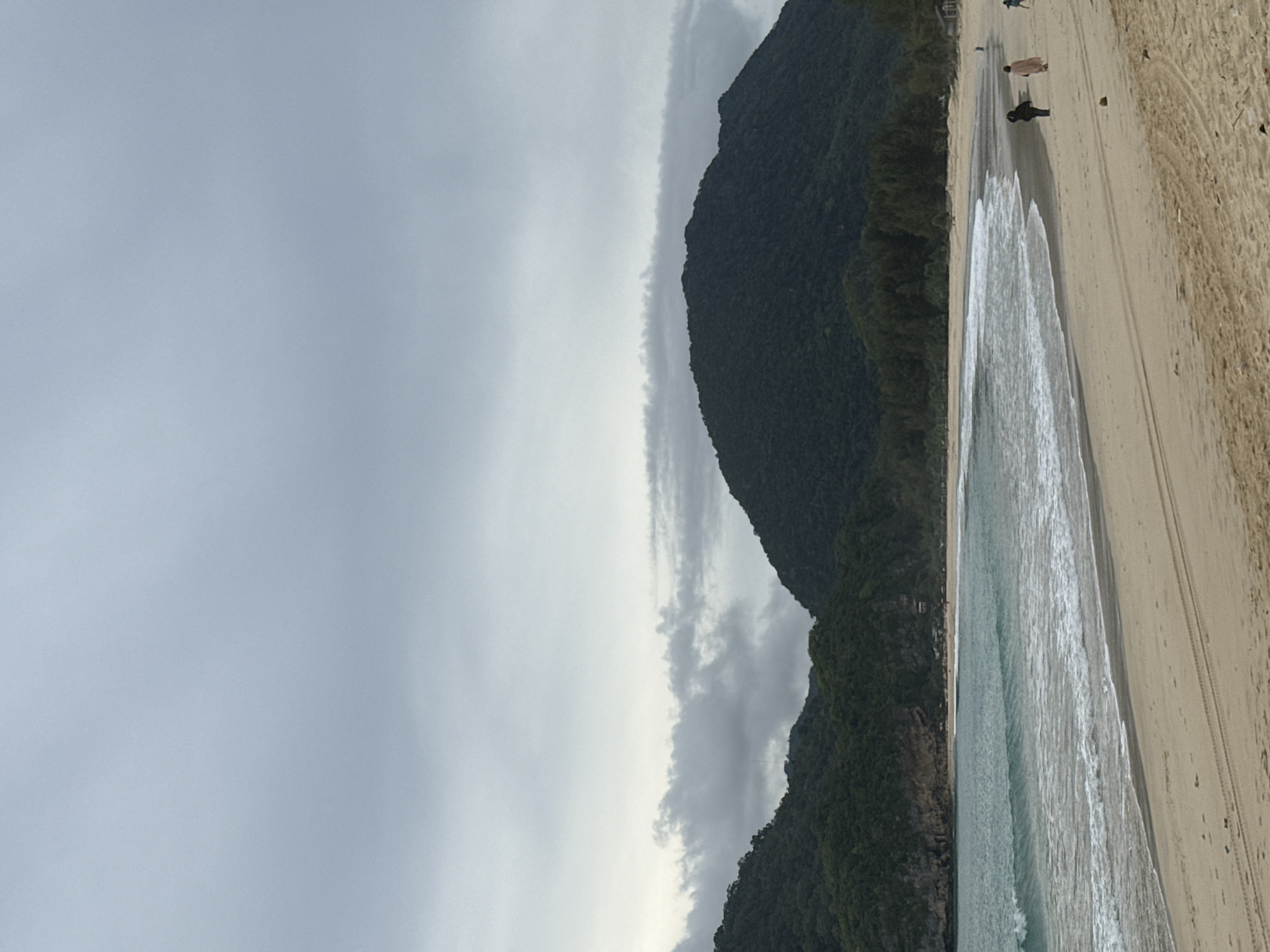 A low-angle, full-shot, portrait-orientation photograph depicts a tropical beach scene under an overcast sky. The scene is divided horizontally, with the left side dominated by a dark, forested mountain that slopes down towards the sea. The right side of the frame features a sandy beach, with gentle waves lapping at the shore. The water is a clear turquoise, and the sand is light brown.
Several people are visible walking along the beach. One person in the foreground appears to be walking towards the left of the frame, while two others are further up the beach, walking in the same direction. The people are small in the frame, indicating a wide expanse of beach and sea.
The weather appears to be cloudy and possibly humid, typical of a tropical climate. The time of day is difficult to ascertain due to the overcast conditions, but the lighting suggests it could be morning or late afternoon. The location cues, such as the lush green mountain meeting the sea, are consistent with a tropical island setting. There are no visible text elements in the image. The overall mood is one of peacefulness and natural beauty.