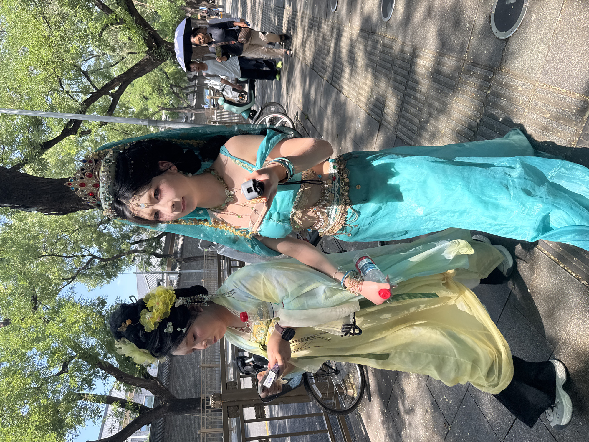 In Xicheng District, China, on a sunlit day, two young women dressed in elaborate, brightly colored costumes are walking on a paved sidewalk. The woman on the left wears a pale yellow, flowing robe with intricate golden embroidery and a tall, elaborate black hairstyle adorned with yellow flowers and pearls. She holds a small, rectangular object, possibly a phone, in her left hand and a bottle with a red cap in her right. The woman on the right is adorned in a vibrant turquoise blue outfit that includes a crown with red and blue jewels, delicate gold chains around her neck and waist, and sheer sleeves. She also has a dark, elaborate hairstyle and is holding a mobile phone in her right hand. Both women are walking from left to right. Behind them, a metal fence borders the sidewalk, and beyond that, trees with lush green foliage line the street. In the background, other pedestrians are visible, including a man with an umbrella and two people walking. A scooter is parked on the other side of the fence. The ground is paved with textured tiles, and shadows from the trees are cast across the scene.