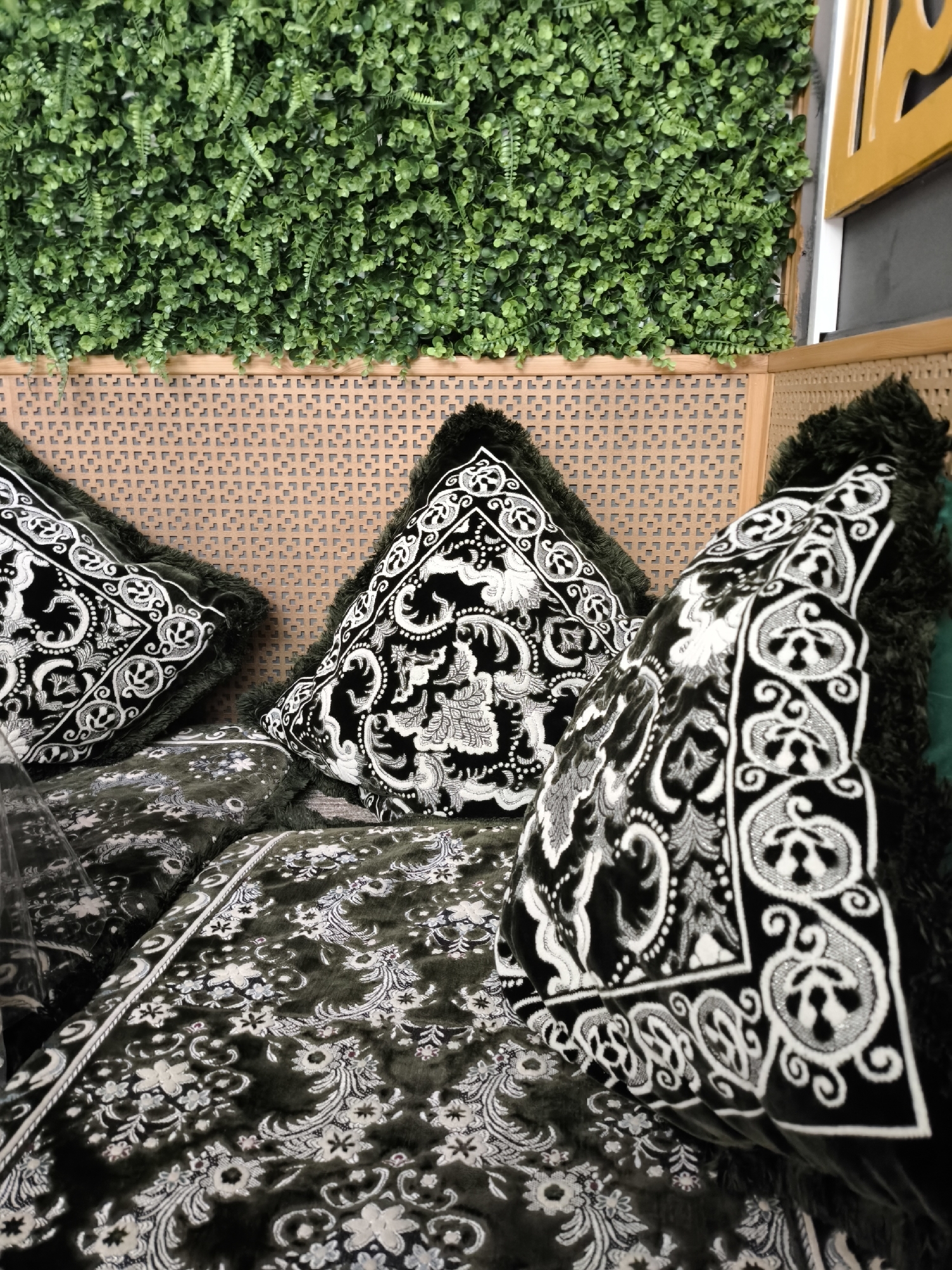 The image depicts a close-up view of an outdoor seating area, likely a patio or balcony. Dominating the foreground are several plush decorative pillows and cushions arranged on what appears to be a sofa or bench. The pillows feature a dark green velvet fabric with intricate white and silver embroidered patterns of floral motifs and swirling designs. The edges of the pillows are trimmed with a thick, dark faux fur.
Behind the seating, a wall is constructed from a light brown, woven material with a repeating geometric pattern. Above this woven panel, a lush green artificial plant wall is installed, creating a vibrant, leafy backdrop. The artificial foliage appears dense and well-maintained, with a mix of small, rounded leaves and some fern-like fronds.
To the right of the frame, a portion of a yellow-lacquered wooden structure is visible, suggesting a partial enclosure or decorative element of the seating area. The lighting suggests it is daytime, with some shadows indicating the presence of natural light. No people are visible in the image.