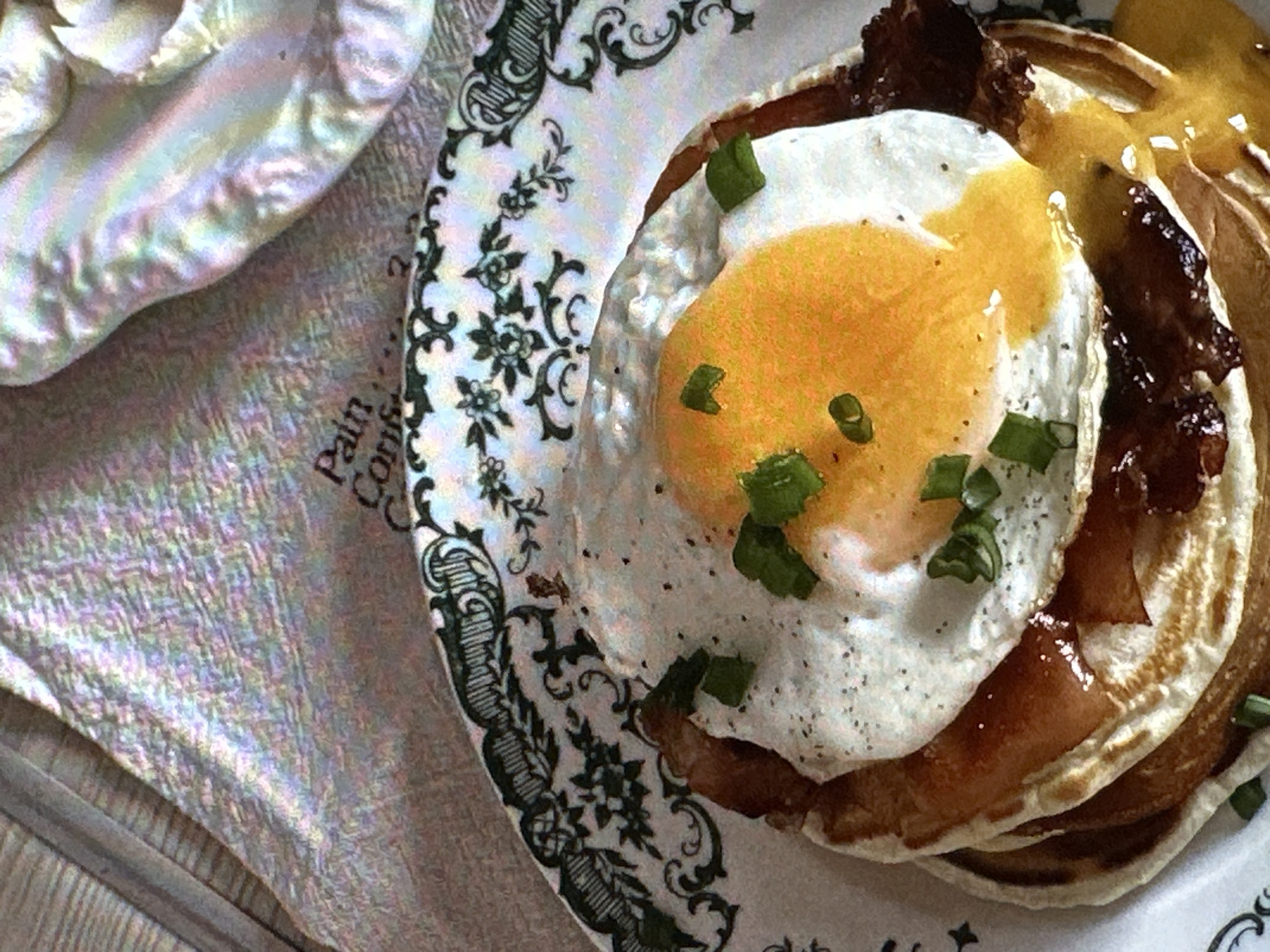 A stack of pancakes topped with a fried egg and bacon is presented on a decorative plate. Chopped green onions garnish the egg. The plate rests on a textured surface beside a fabric-covered object. The location is not identifiable from this image.