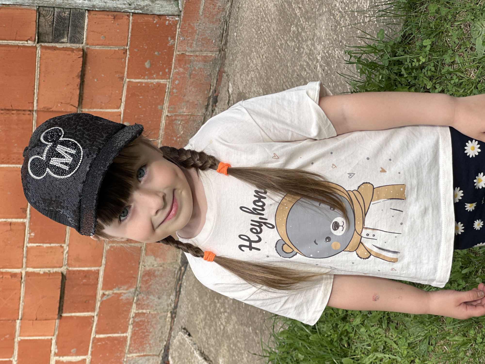 A young girl stands outdoors, her body angled slightly to the right. She wears a black, sequined baseball cap with a white Mickey Mouse emblem. Her light brown hair is styled in pigtails secured with orange ties. She has fair skin and blue eyes. She is wearing a white t-shirt with a cartoon bear design. The background consists of a red brick wall on the left, a concrete surface, and green grass and vegetation on the right. Based on visual cues, the image does not provide definitive evidence to confirm the location in Chaikovsky, Russia.