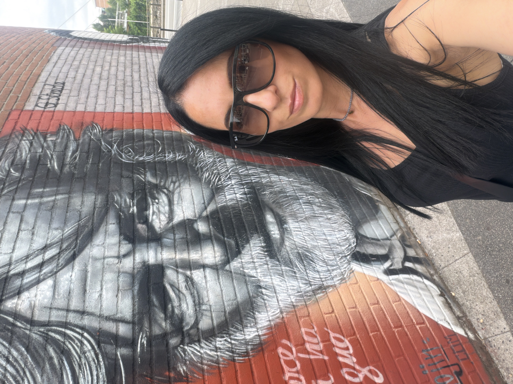 In Corsico, Italy, a woman with long black hair and sunglasses stands in front of a large mural on a brick wall. The mural is a black and white portrait of an elderly man with a beard, painted over a red brick background. There is some text in white painted onto the red part of the wall. The woman is wearing a black sleeveless top and a necklace. She is looking towards the camera, taking a selfie. The environment appears to be outdoors, with a sidewalk and a street visible in the background.