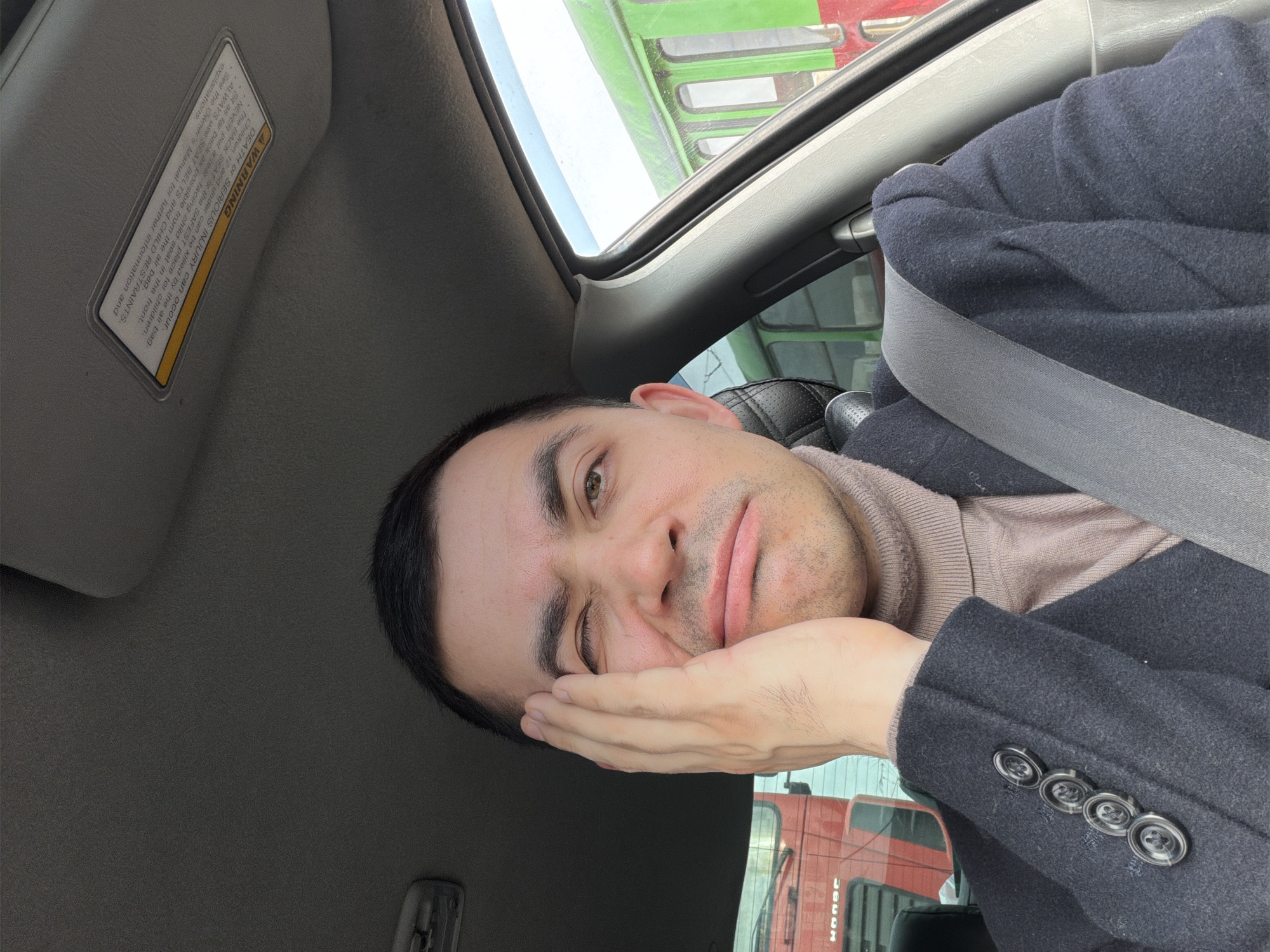 The image shows a man in a car, wearing a dark coat with four buttons on the sleeve and a light-colored turtleneck sweater. He has dark hair, brown eyes, and is resting his face in his hand with a slightly weary expression. The car's interior is visible, including a sun visor with a warning label, a sunroof, and a seatbelt. Through the window, parts of a green and a red truck are visible, with Cyrillic text on the red truck. Based on the Cyrillic script, the location is likely in Russia, and the trucks visible are suggestive of a commercial area. Since I was asked to provide information about the location in Izhevsk, Russia, I will add that the presence of commercial vehicles may indicate proximity to an industrial or transport area within that city.