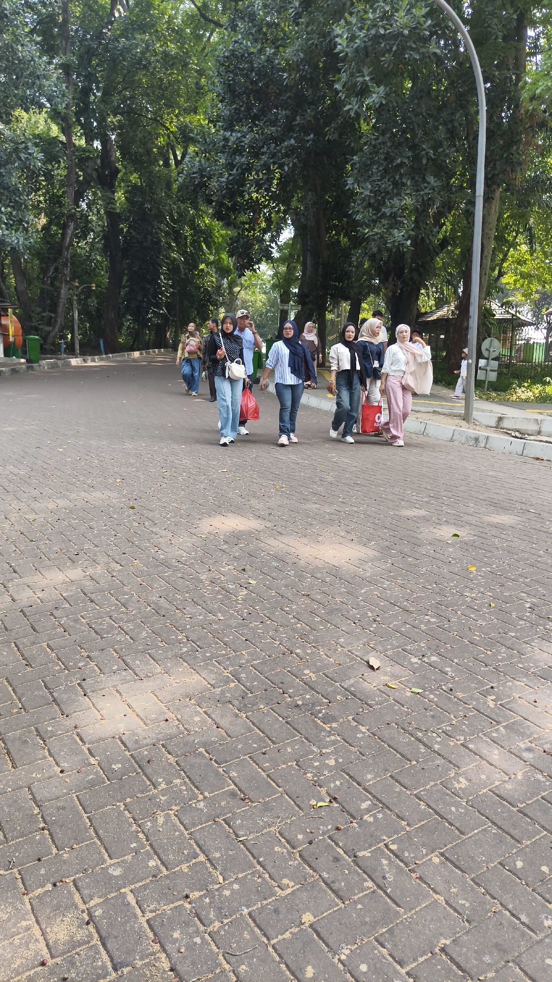 The image depicts a group of people walking on a paved walkway in a park-like setting. The walkway is made of interlocking grey pavers and is lined with trees. In the background, there is a street and a fence. The people are dressed in casual clothing, and some are carrying bags. One person is wearing a blue and white striped shirt and jeans, another is in a white shirt and pink pants, and a third is in a black and white patterned top and jeans. The overall scene suggests a leisurely day in a public space. The location is identified as Gelora Bung Karno (GBK) Sports Complex, Central Jakarta, Indonesia.