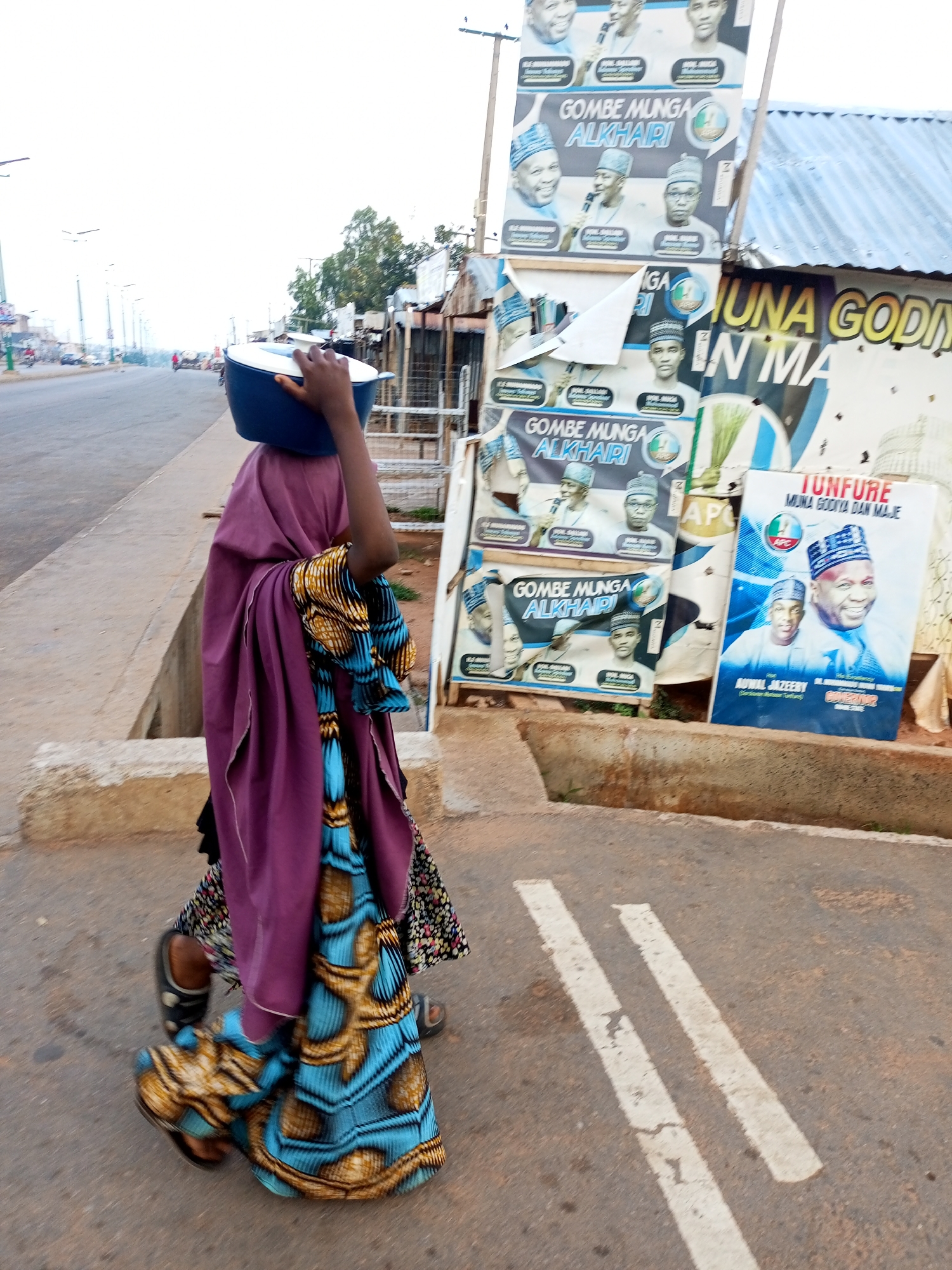 In Gombe, Nigeria, a young person wearing a purple hijab and a brightly patterned blue and gold skirt walks along a paved road, balancing a dark blue basin on their head. The person is walking away from the camera, towards a street lined with stalls and posters. The posters, some torn and faded, advertise political candidates, with prominent images of men in traditional caps. The street is bordered by a raised concrete curb and a narrow drainage ditch. In the background, trees are visible, and a few vehicles are on the road, suggesting the bustling nature of the city. The overall scene captures a slice of everyday life in Gombe, with a blend of commerce, politics, and pedestrian activity.