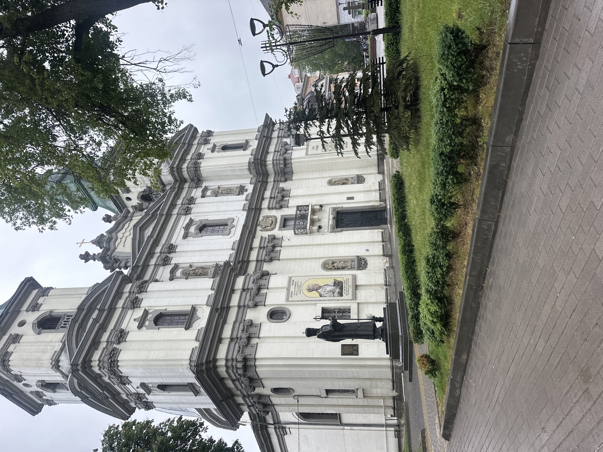 The image displays an ornate Baroque-style church facade in Ternopil, Ukraine, under an overcast sky. The large, multi-story building features a light-colored exterior with prominent dark grey architectural details, including cornices, pilasters, and window frames. The facade is richly decorated with sculptures housed in niches and various decorative friezes.
A notable feature on the church's exterior is a rectangular mosaic panel depicting Pope John Paul II, identifiable by his attire and the clearly legible dates "1920 2005" below his image, commemorating his life. Directly in front of this mosaic, a dark-colored statue of a robed religious figure, possibly a bishop or pope, stands on a stone pedestal, holding a staff.
To the left of the church, a large deciduous tree with dense green foliage partially obscures the upper portion of the building. To the right, a paved sidewalk made of grey interlocking pavers runs alongside a strip of well-maintained green grass and neatly trimmed bushes. A tall, decorative black lamppost with multiple light fixtures is visible further to the right. In the background, glimpses of other urban buildings can be seen beyond the lamppost and trees. The scene is static, capturing the architectural beauty of the church within its urban context on a cloudy day.