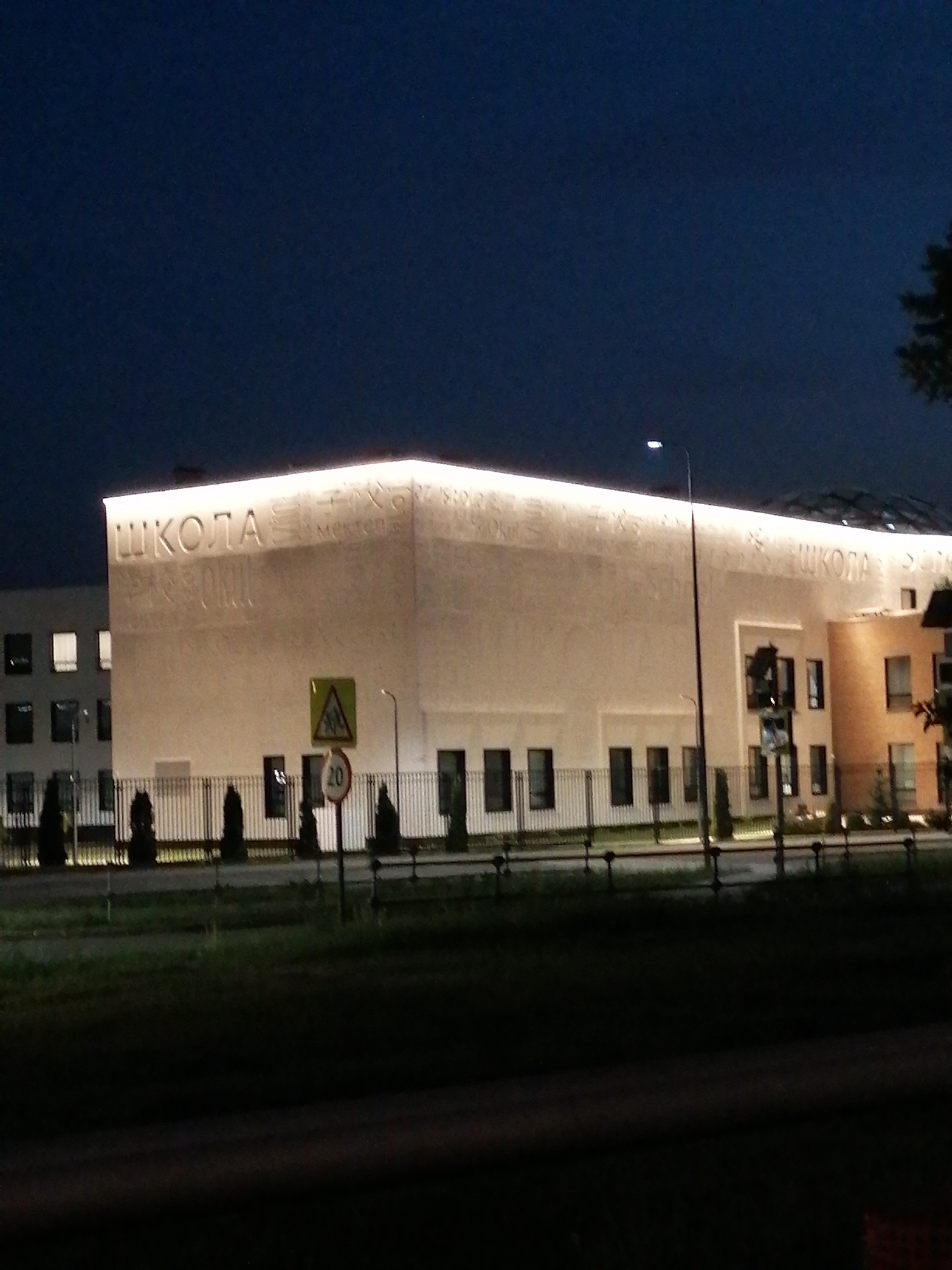 The image depicts a building at night, likely a school, as suggested by the Russian word "ШКОЛА" prominently displayed on its facade. The building is illuminated with horizontal lights along its top edge, and there are other lights shining on the facade. Several windows are visible on the side of the building. In front of the building is a fenced area with some trees or shrubs. Street signs, including a triangular pedestrian crossing sign and a speed limit sign indicating 20, are visible near the fence. A streetlight and traffic lights are also present near the building. The location is Unknown, Unknown. The scene appears calm and orderly.