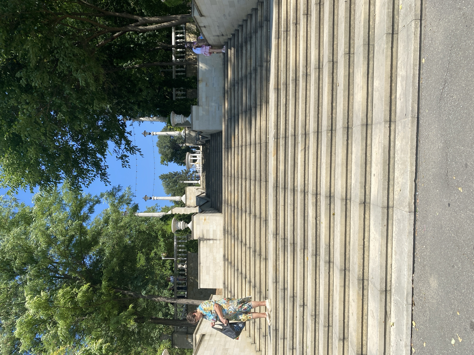 The image captures a wide, imposing stone staircase leading upwards towards a park-like setting with lush green trees and clear blue skies. The architecture suggests a public space, possibly a monument or a grand entrance to a park or cultural institution. The staircase is constructed from large, light-colored stone blocks, with multiple landings and a symmetrical design.
On the left side of the lower part of the staircase, a woman in a brightly colored floral dress is standing, looking down at a dark tote bag she is holding. She is wearing light-colored shoes. Her posture and the way she holds the bag suggest she might be paused or about to descend.
Further up the staircase, on the right side, two young girls in light-colored clothing are visible. One, wearing a pink skirt and white top, appears to be climbing the stairs, while the other, behind her, is partially obscured.
The environment is sunny, with shadows cast by the trees and the architecture on the stone steps, indicating a midday or early afternoon time. The presence of trees, planters, and decorative urns on the upper levels suggests a well-maintained outdoor public space.
This staircase is a prominent feature of the Alley of Classics in Chișinău, Moldova. The Alley of Classics is a popular park and promenade located in the center of Chișinău, lined with busts of prominent Moldovan writers and cultural figures. The grand staircase leading up to the park is a well-known landmark, offering panoramic views of the city from its upper levels. The setting is consistent with the architectural style and landscaping found in this area of Chișinău.