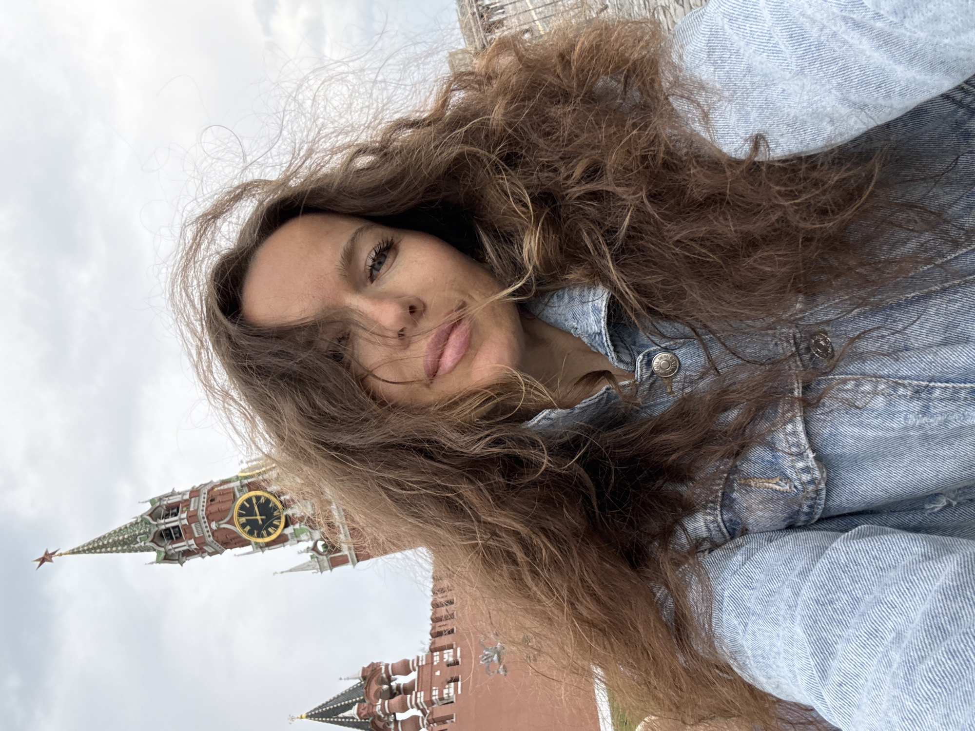 A woman with long, wavy brown hair and blue eyes is taking a selfie in front of the Spasskaya Tower of the Moscow Kremlin. She is wearing a light blue denim jacket. The Spasskaya Tower, a prominent landmark of the Kremlin, is visible in the background with its red brick walls and a large golden clock. The sky is overcast.