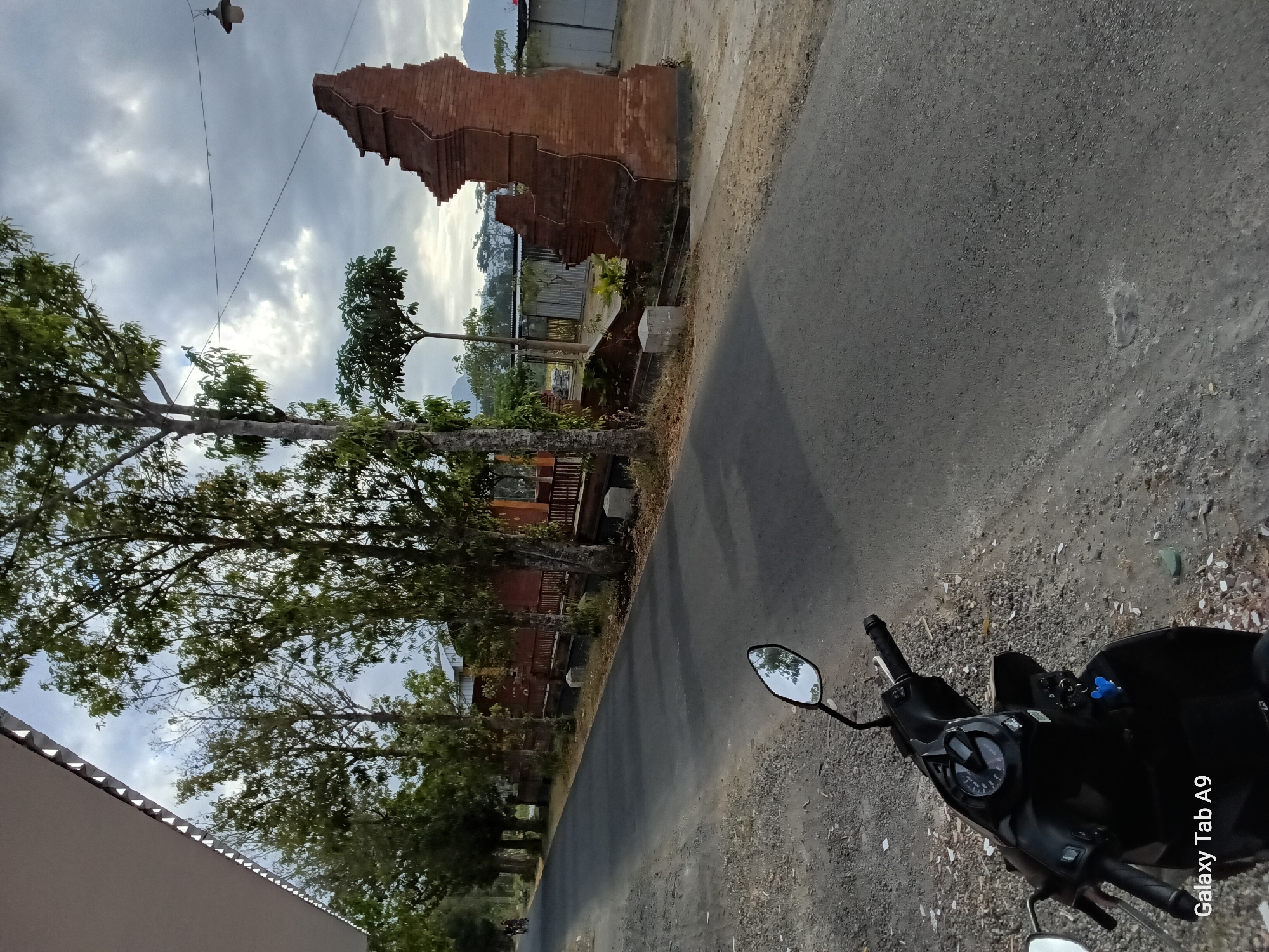 The image depicts a street scene in Bendorejo, Indonesia, under an overcast sky. In the foreground, a black scooter is parked on the gravel shoulder of an asphalt road. The scooter's side mirror reflects trees and the sky. Along the right side of the road, a weathered, brick gateway structure, possibly a temple ruin or architectural element, stands prominently. Beyond the gateway, there are buildings and vegetation. A line of tall trees with sparse foliage flanks the left side of the road, leading into the distance. In the background, mountains are faintly visible. The overall atmosphere is subdued, suggesting an early morning or late afternoon.