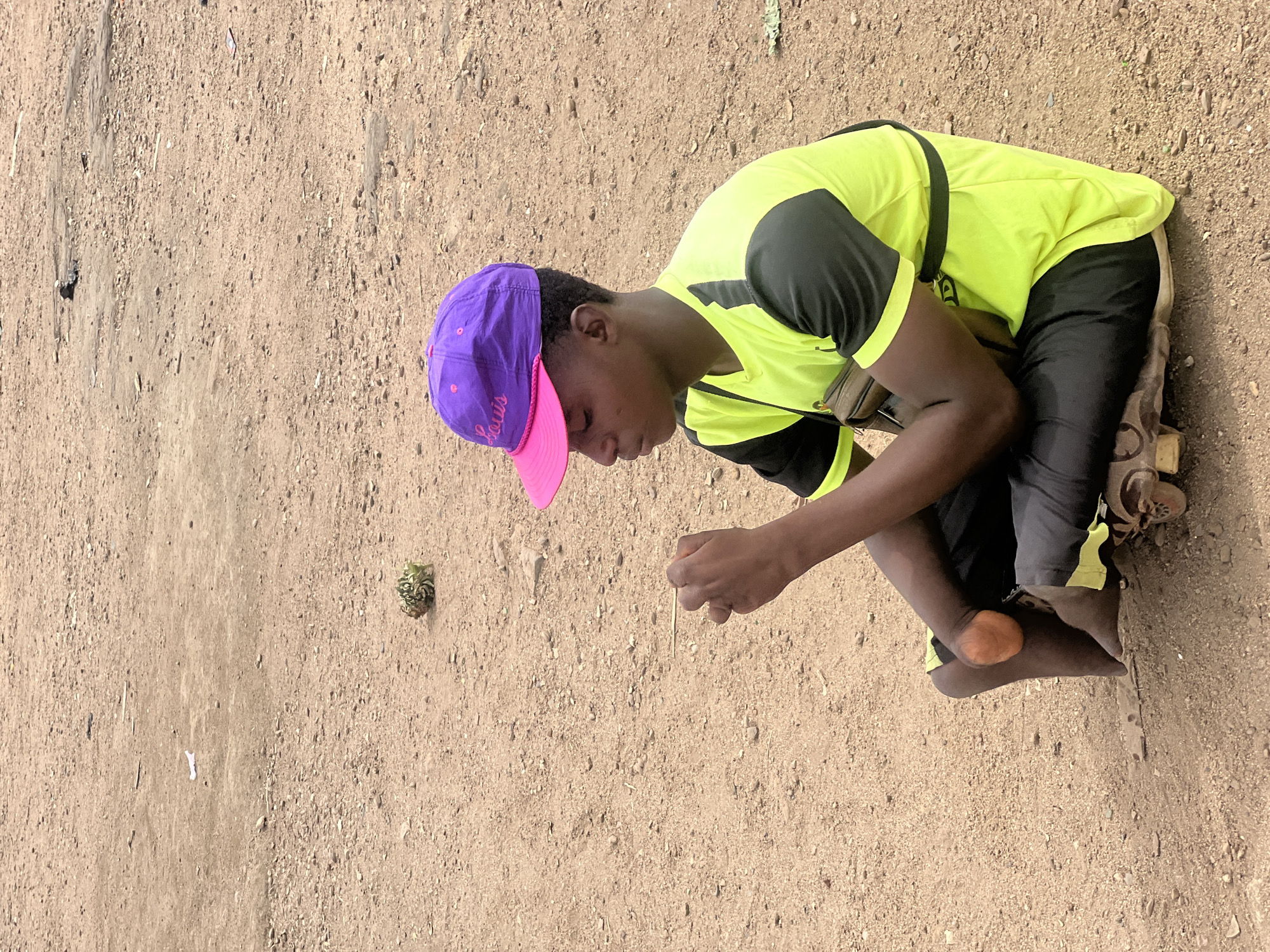 A young man kneels on the ground in Bukuru, Nigeria. He wears a purple and pink baseball cap, a neon green and black athletic shirt, and black pants. He holds a small stick in his right hand and appears to be examining a thorny fruit-like object on the ground to his left. The ground is a mix of tan sand and small pebbles.