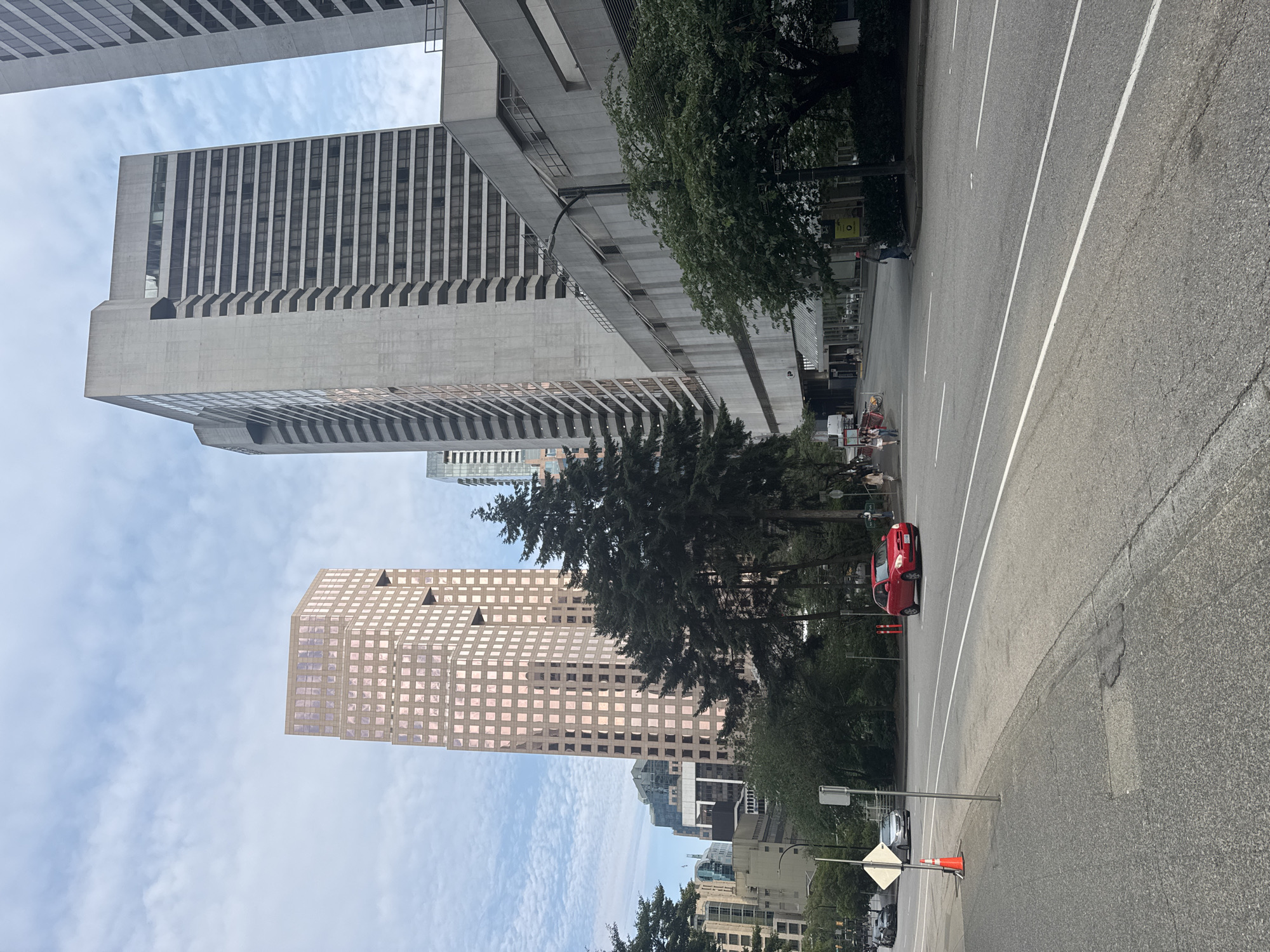 The image shows a street view in Vancouver, Canada, with several tall buildings and trees. In the foreground, a gray asphalt road with white lane markings curves to the right. A red car is driving on the road. To the left of the road, there is a sidewalk, a traffic cone, and a street sign. In the background, there are several modern skyscrapers. One prominent building is a tall, octagonal structure with a pinkish-brown facade and a grid of windows. Another is a tall, gray building with a striped facade. Dense evergreen trees are interspersed between the buildings and the road. The sky is partly cloudy. People are visible walking on the sidewalk and in the distance.
