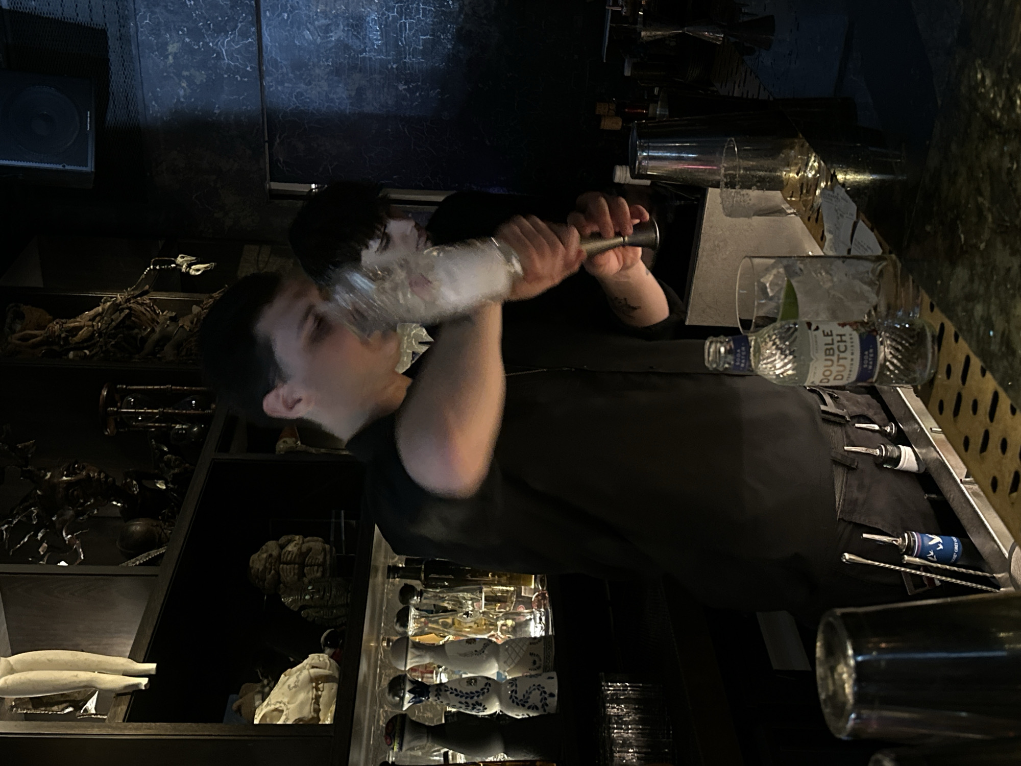 The image captures an evening scene inside a dimly lit bar, likely in London, United Kingdom. Two bartenders are actively preparing drinks behind a dark counter.
The primary subject is a man in the foreground, wearing a dark short-sleeved shirt, who is vigorously shaking a cocktail shaker. His arm and the shaker are blurred due to motion, indicating the intensity of the action. He looks focused on his task. Behind him, another person, also wearing a dark top, is partially visible and appears to be holding another shaker or bar tool, suggesting a busy bar environment.
The bar counter features various tools and glassware. A tall glass filled with ice stands prominently, next to a small, clear glass bottle labeled "DOUBLE DUTCH" with "SODA WATER" and "Premium Mixers" clearly visible. Other bar essentials like jiggers, pour spouts, and possibly stirrers are neatly arranged in recesses on the counter. Below the counter, a display of ornate, possibly ceramic, bottles or dispensers can be seen.
The background is adorned with an eclectic mix of decorative items displayed on dark shelves. Notable objects include a large speaker, various skeletal or bone-like sculptures (including what looks like a dinosaur skeleton piece and a skull), mannequin legs, an hourglass, and a brain-like sculpture. The overall lighting is low, creating a sophisticated and somewhat mysterious atmosphere, typical of an upscale bar or lounge during the evening hours.