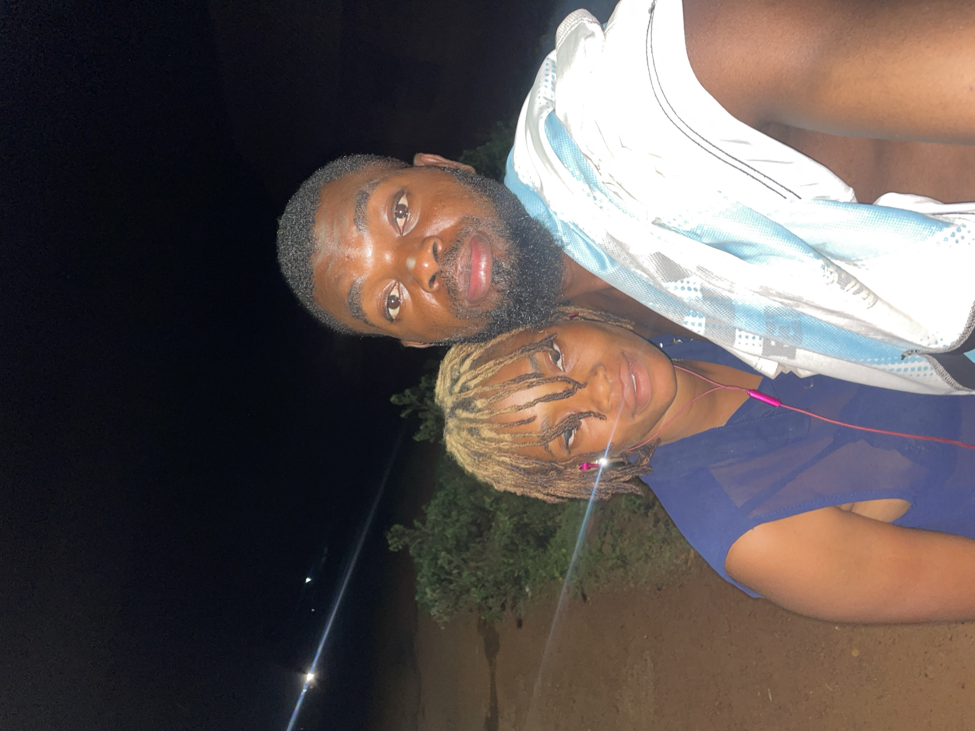 A man and woman are in a selfie photograph taken outdoors at night in Ogbe, Nigeria. The man has a beard, and he is wearing a white garment. The woman has blond dreadlocks and is wearing a blue top. Pink earphones are visible around her neck. Trees are visible in the background.