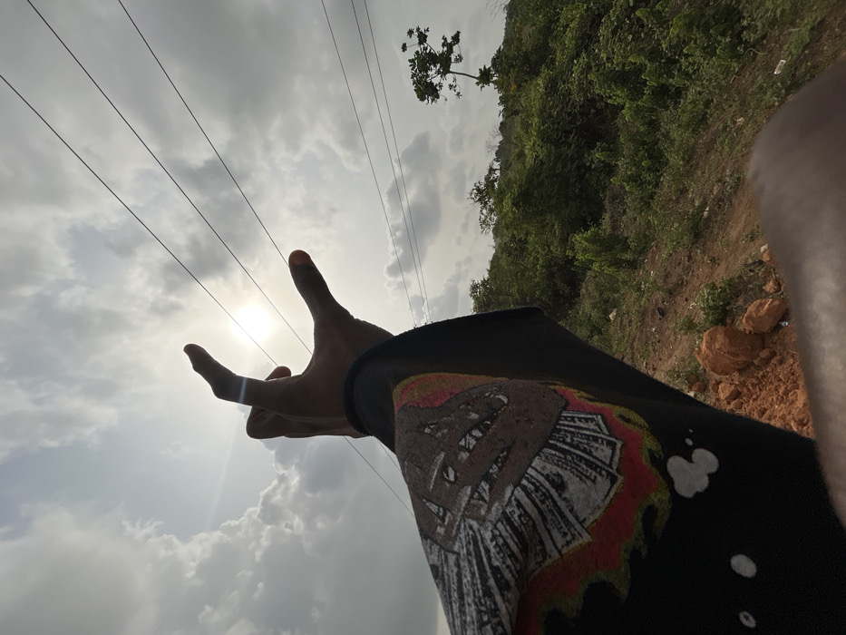 The image depicts a low-angle outdoor scene in Ilishan, Nigeria, viewed upwards towards the sky and horizon. In the immediate foreground, a dark-skinned human left arm, covered by a black long-sleeved garment, extends towards the upper center. The hand is positioned with fingers slightly curved, appearing to "hold" or "pinch" the bright, partially obscured sun between the thumb and forefinger. The garment's sleeve features a graphic design comprising a skull-like image made of what appears to be fanned dollar bills, accented with red and white elements, possibly resembling flames, on a black background.
The sky occupies the upper two-thirds of the frame, characterized by significant white and grey cloud cover. The sun, a bright circular disc, is centrally positioned, emitting a lens flare. Three dark, parallel power lines traverse the sky horizontally in the middle ground, extending from the left past the hand towards the right.
The background, predominantly in the lower right quadrant, reveals a lush, green, hilly landscape. This terrain is densely covered with various types of foliage and bushes. A distinct, sparsely leafed tree stands vertically on the far right against the sky. Patches of reddish-brown earth and scattered rocks are visible in the foreground along the bottom and lower right, suggesting bare ground or a roadside.