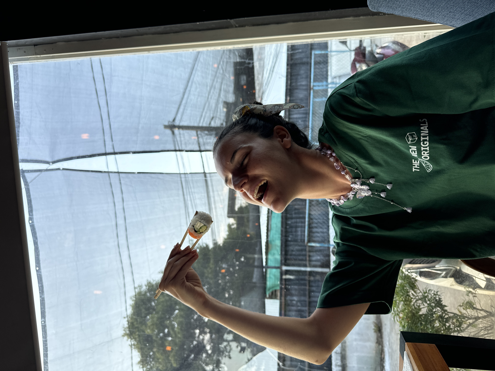 A woman is holding a piece of sushi with chopsticks over her head, her mouth open in laughter. She is wearing a green t-shirt with a logo that reads "THE NEW ORIGINALS" and a necklace with purple beads and butterfly charms. The scene appears to be indoors, by a window that looks out onto a street with trees and power lines. The window is covered with a sheer white curtain that diffuses the light. In the background, there are glimpses of buildings and vehicles. The lighting suggests it is daytime. The overall mood is playful and joyous.
