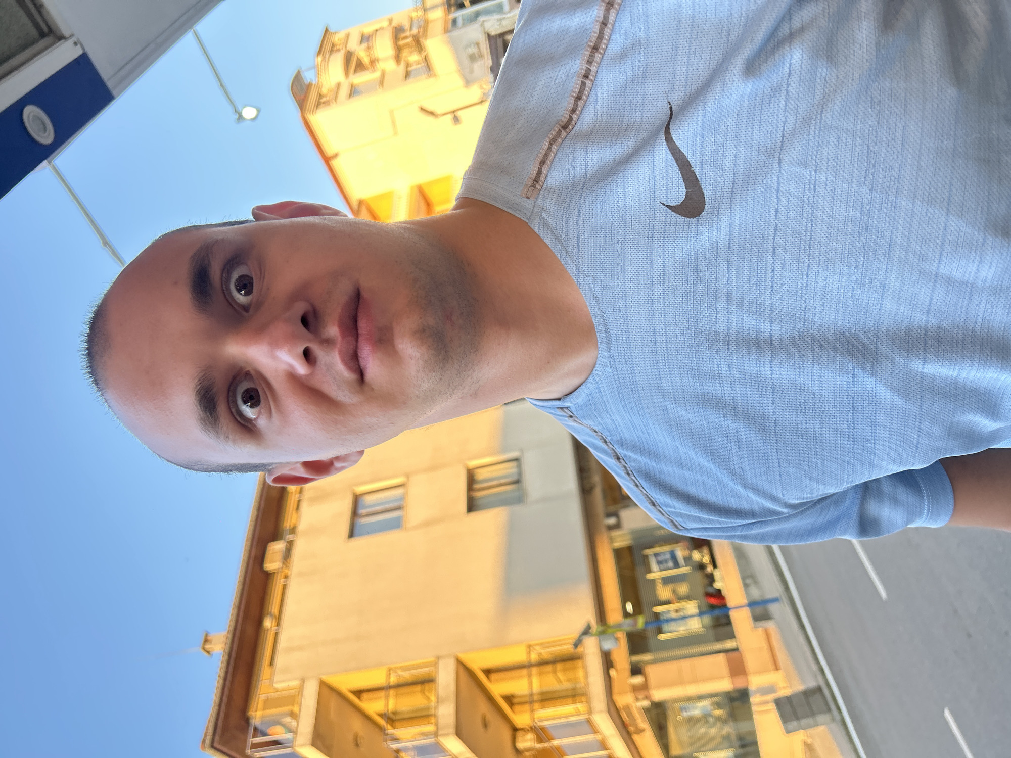 A man is standing in Sozopol, Bulgaria, wearing a light blue Nike t-shirt. He has a bald head and brown eyes, and he is looking up at the camera. Behind him is a golden-colored building with multiple windows and balconies. The sky is clear and blue. A streetlight is visible in the upper left corner.