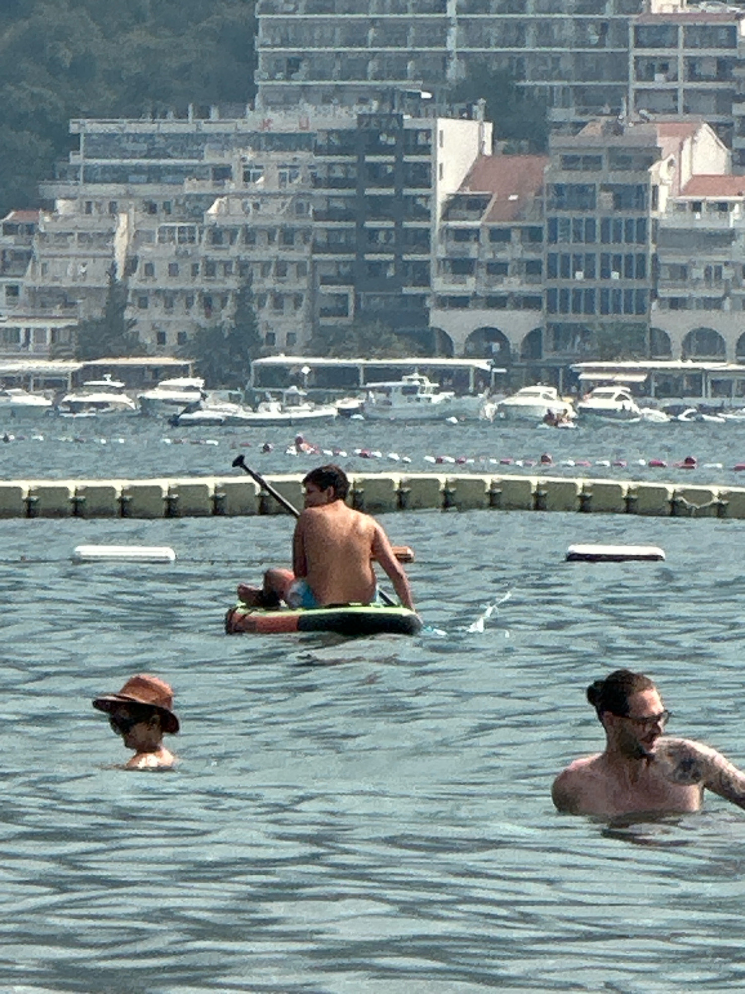 In Boreti, Montenegro, a body of water is visible with several people engaged in aquatic activities. In the foreground, a person wearing a hat and sunglasses floats in the water. To their right, another person, shirtless and with glasses, is also in the water. In the mid-ground, a person is on a paddleboard, facing away from the camera and holding a paddle. Behind them, a yellow barrier with red and white buoys separates the swimming area from other parts of the water. In the background, various buildings, possibly hotels or apartments, are visible along the coastline, with trees and greenery behind them. Several boats are also present in the water.